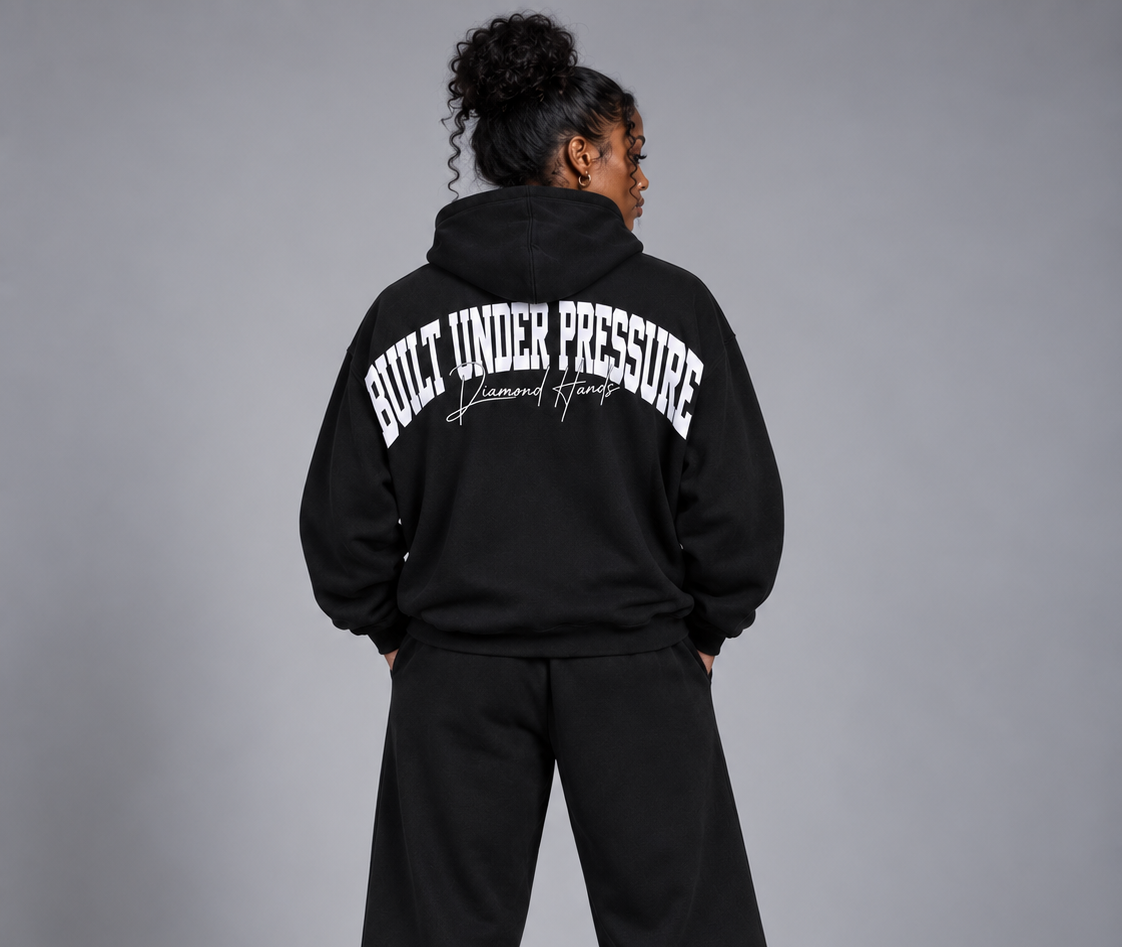 Back view of a woman wearing a black hoodie with white text that says 'Built Under Pressure' and 'Diamond Hands', with her hands in her pockets, standing against a plain gray background.