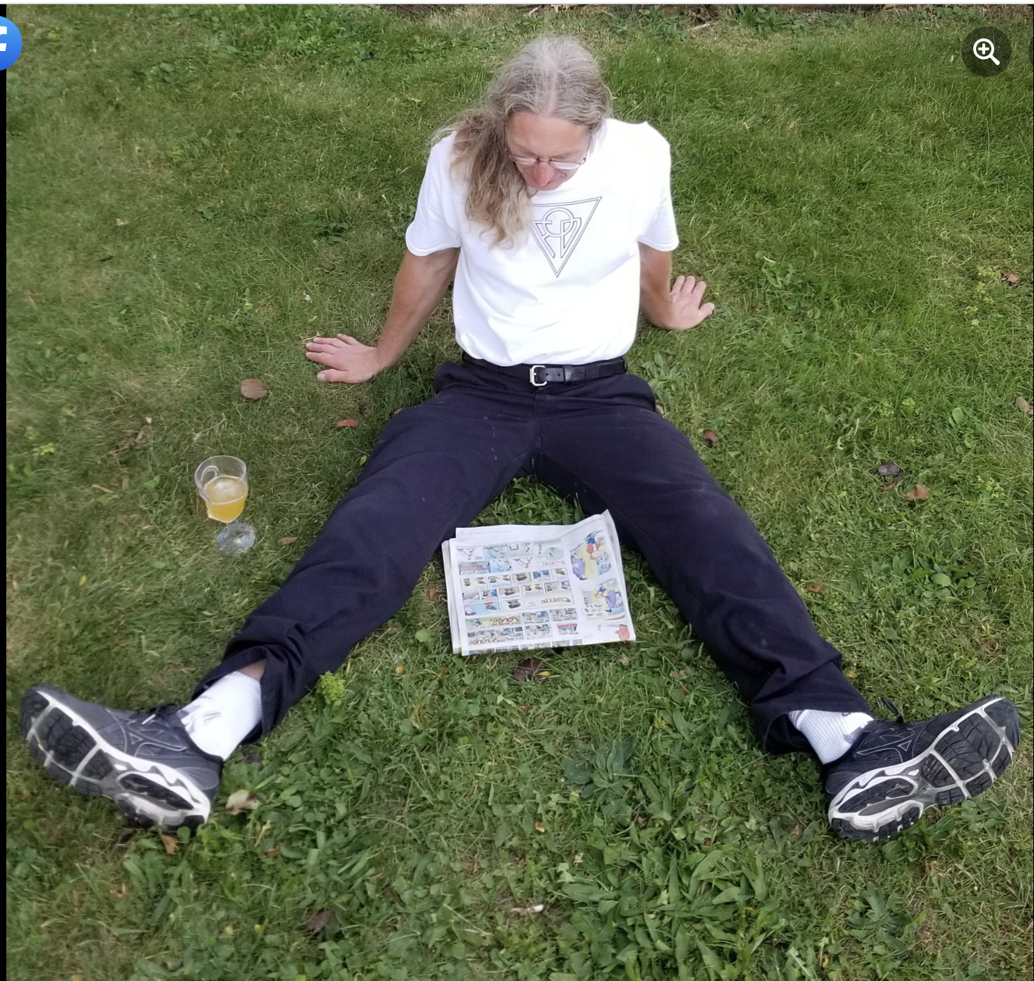 A man with long gray hair and glasses is sitting on the grass with legs spread apart. He is wearing a white T-shirt with a geometric design, black pants, and sneakers. There is a comic book open on the grass in front of him and a glass of beer nearby. The man is looking down at the comic book.
