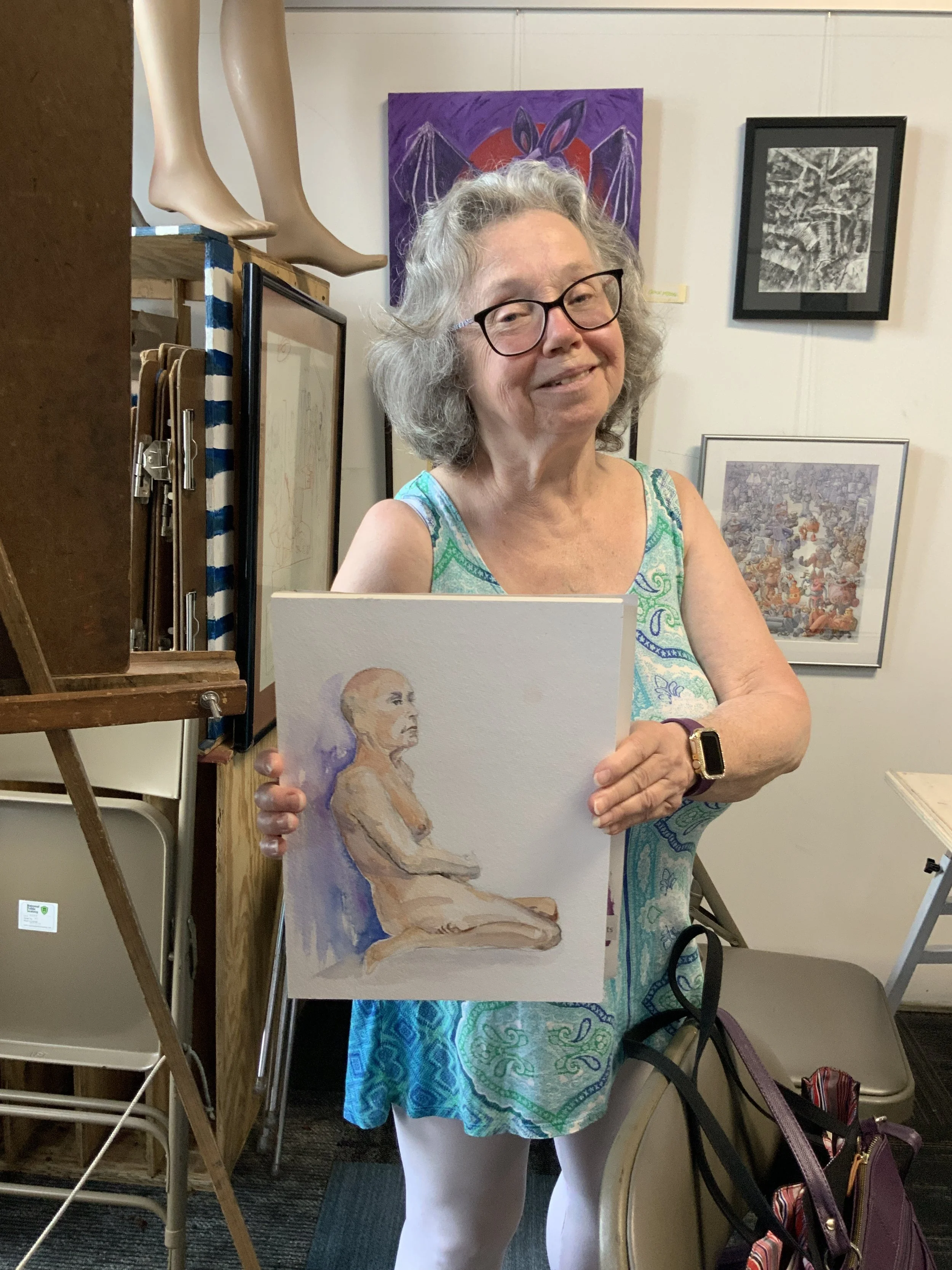 A woman with gray hair and glasses holding a portrait of an older woman in a sitting pose. She is wearing a blue and green patterned dress, standing in an art gallery with various pieces of art on the wall behind her.