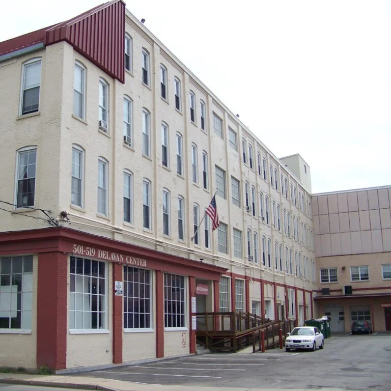 A multi-story brick building with a red trim, labeled as Delavan Center, with an American flag hanging, and a small parking lot in front with a white car and some green trash bins.