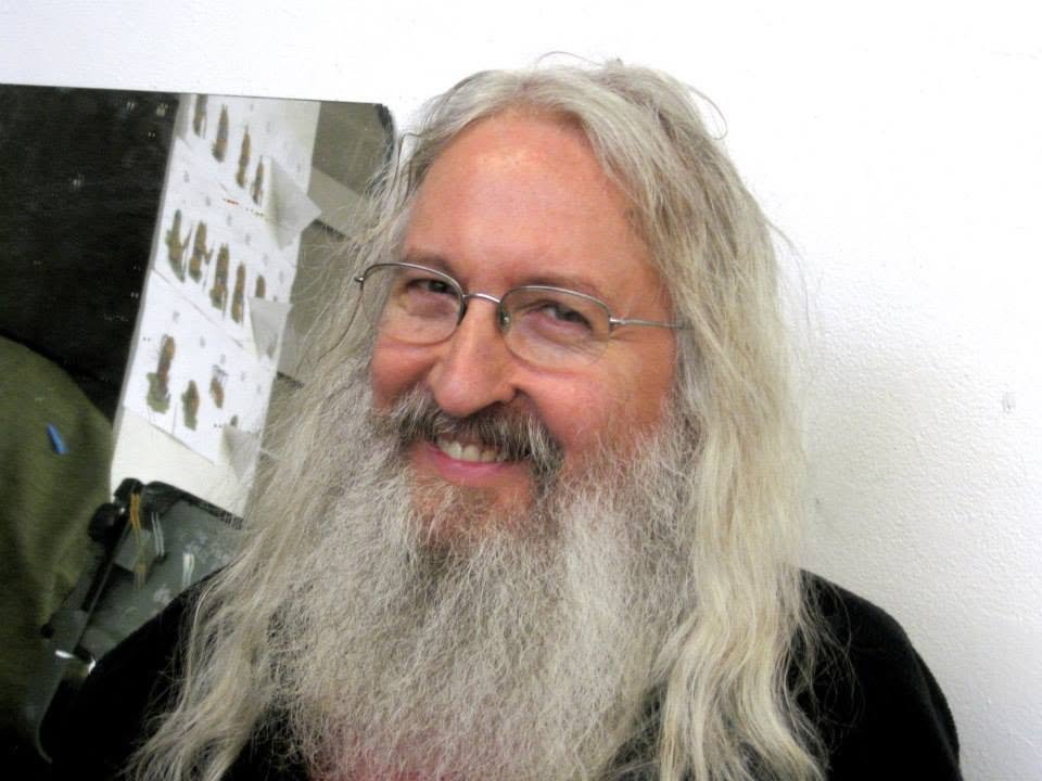 A cheerful man with long gray hair, a beard, and glasses smiling at the camera.