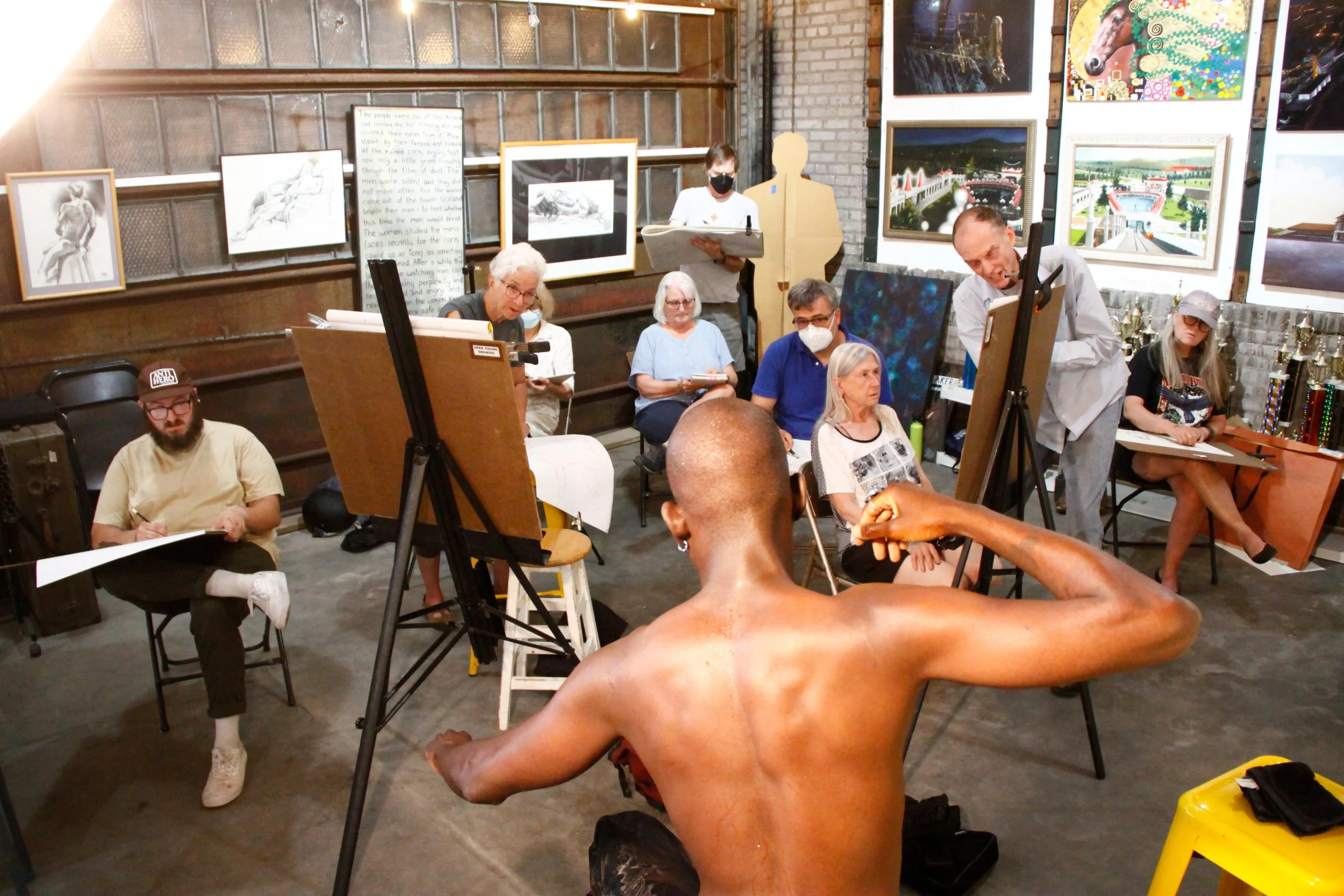 An art class with people working on sculptures and paintings in an industrial-style gallery space. Artworks and a handwritten board are on the walls.