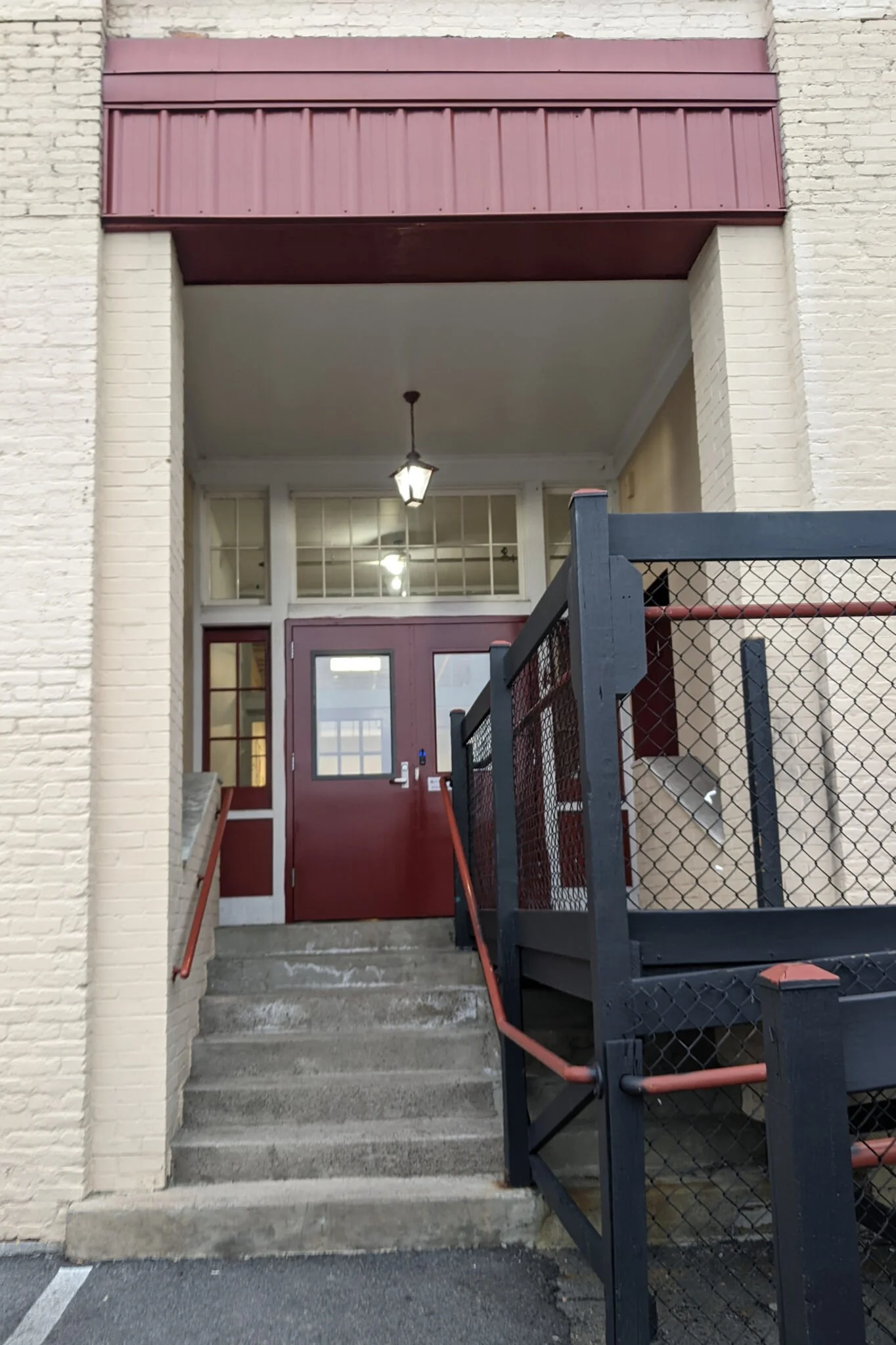 Concrete stairs leading up to a red door with white framing. There is a black metal fence and gate in front of the stairs. The building has white brick walls and a red roof extension with a hanging lantern light.