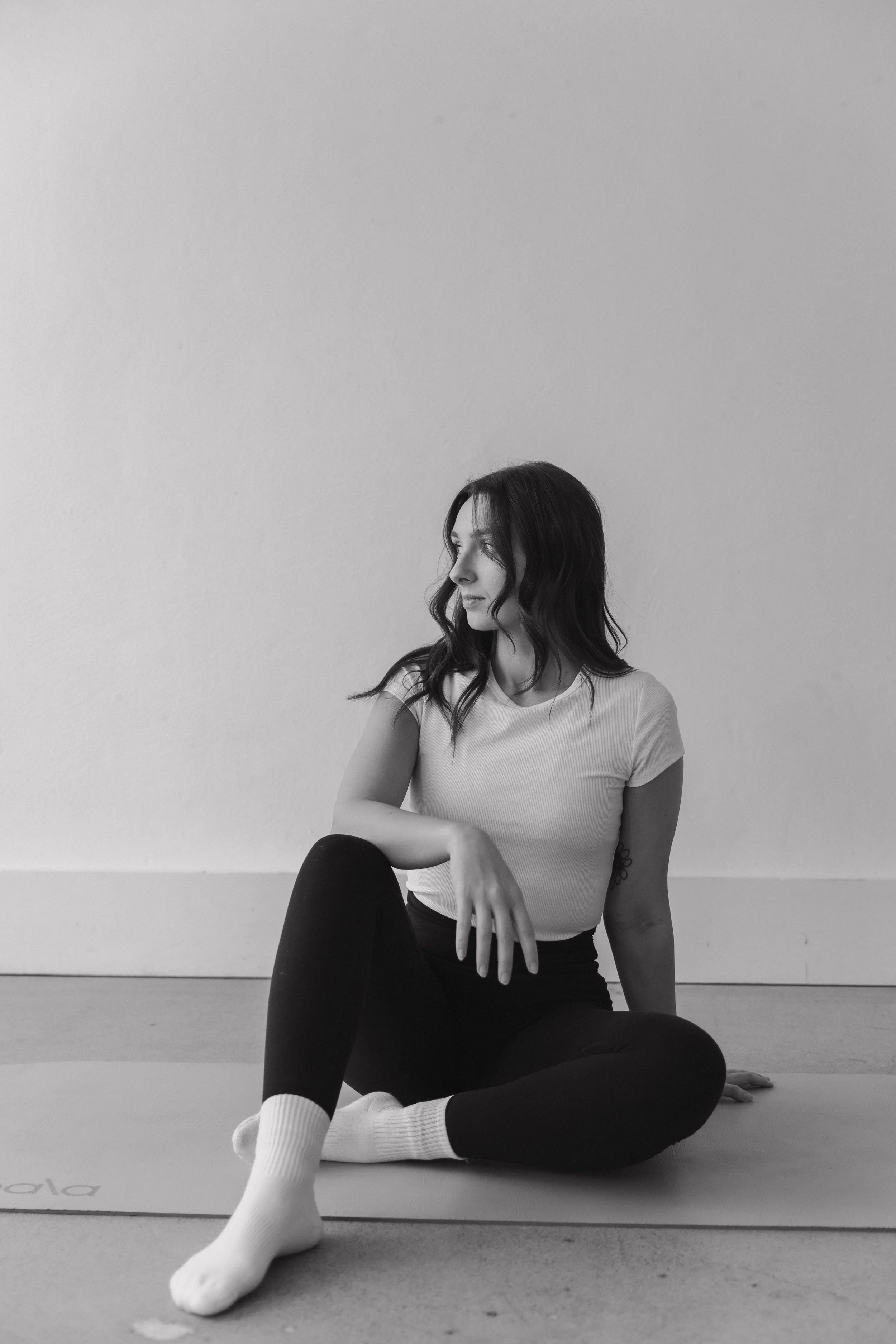 A woman sitting on the floor with her legs crossed, wearing a white t-shirt, black leggings, and white socks, looking to her right against a plain wall.
