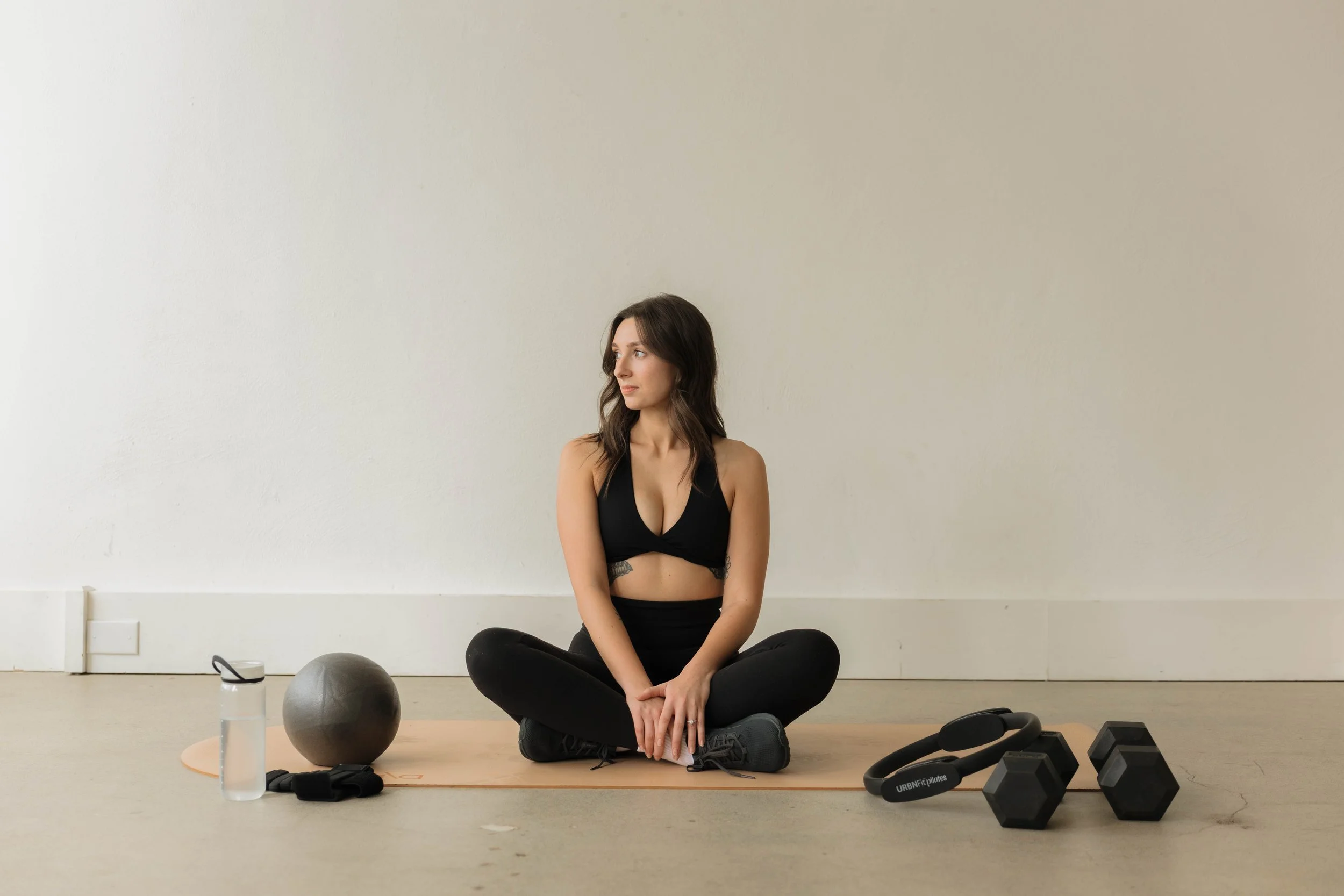 A woman sitting in a cross-legged yoga pose on a light-colored yoga mat in a room with a plain beige wall, surrounded by fitness equipment including a black exercise ball, dumbbells, water bottle, and resistance bands.