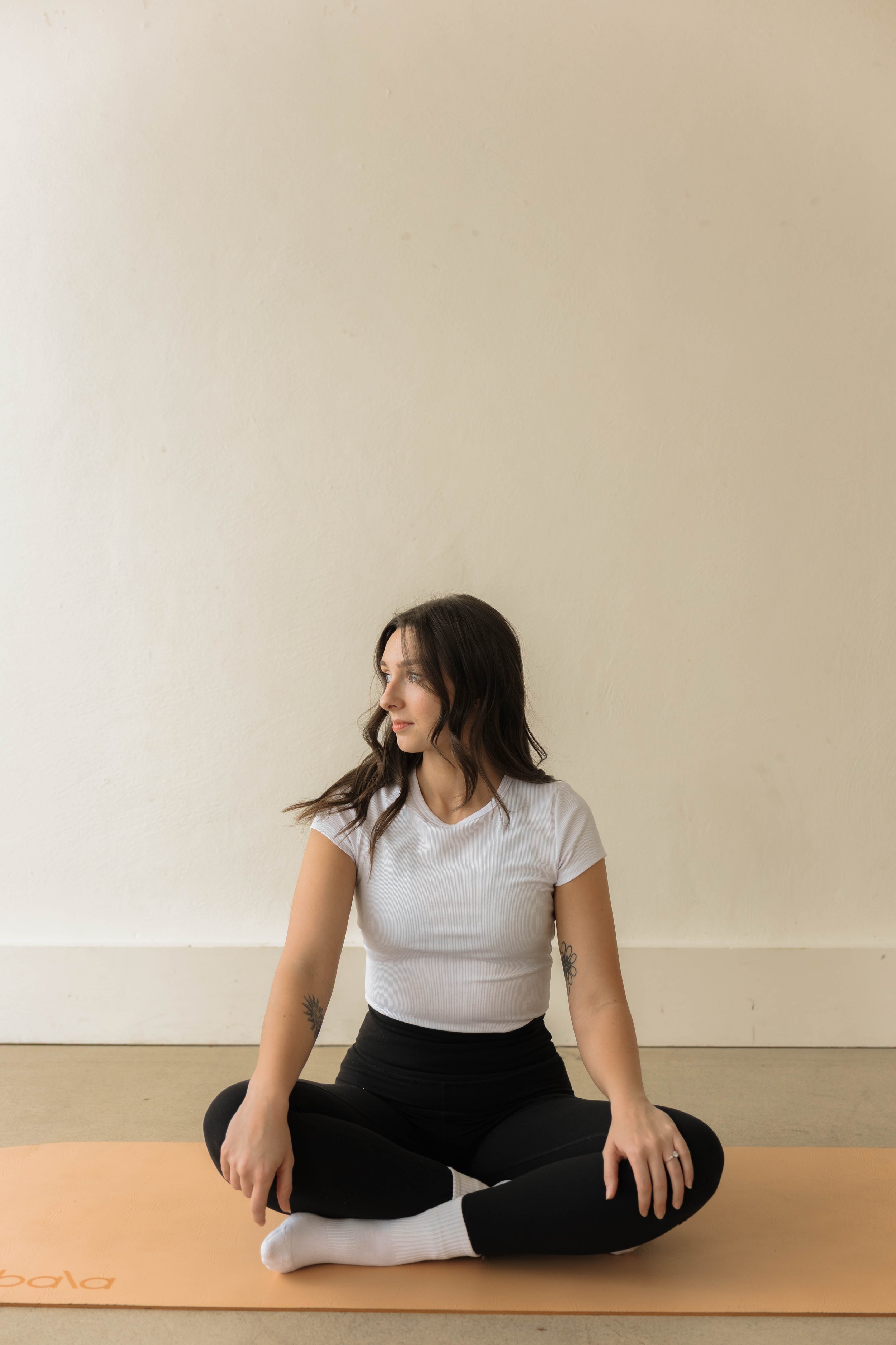 A young woman lined up cross-legged on a yoga mat, sitting in a meditative pose with calm expression, wearing a white t-shirt and black pants, with tattoos on her arms, against a plain beige wall.