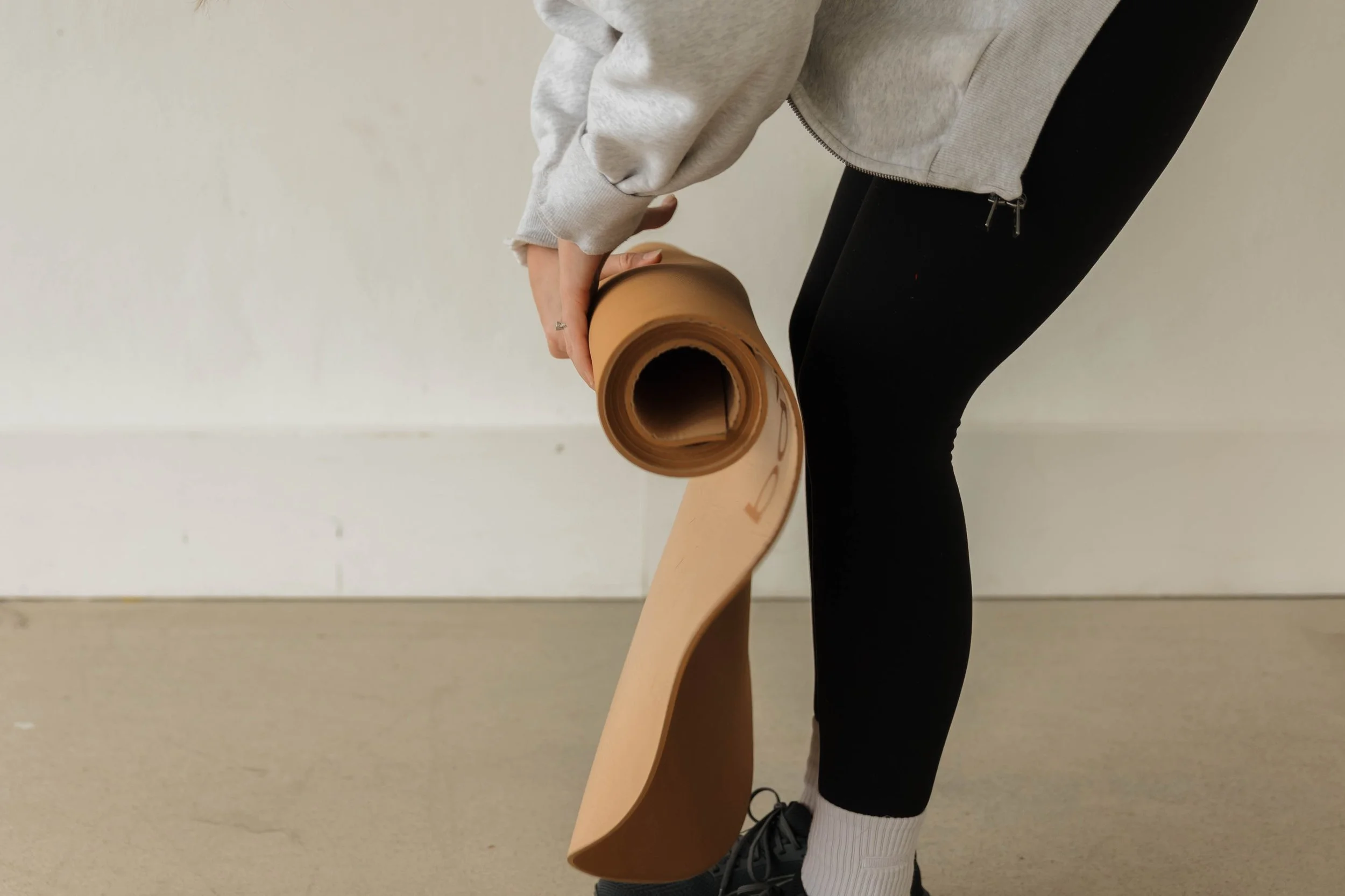 Person rolling up a yoga mat wearing black pants, black qsneakers, and a gray zip-up hoodie.