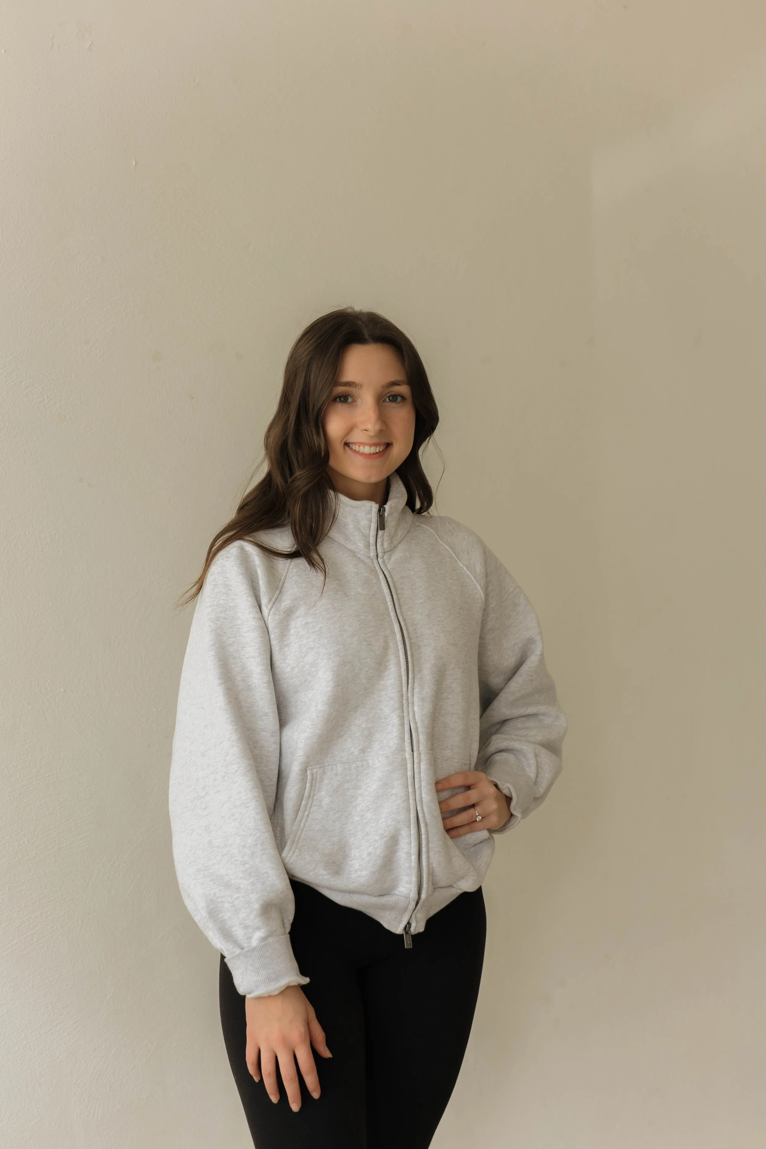A young woman with brown hair smiling, wearing a light gray zip-up hoodie and black pants, standing against a plain off-white wall.