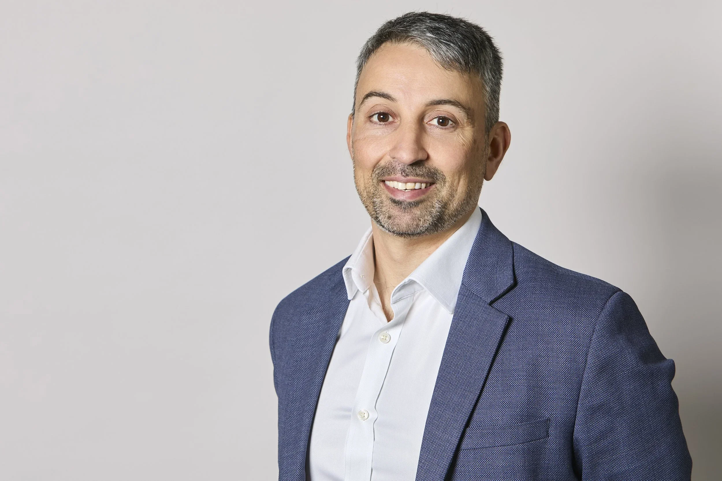 Damien Colella in a white shirt and navy blue blazer against a plain light gray background.