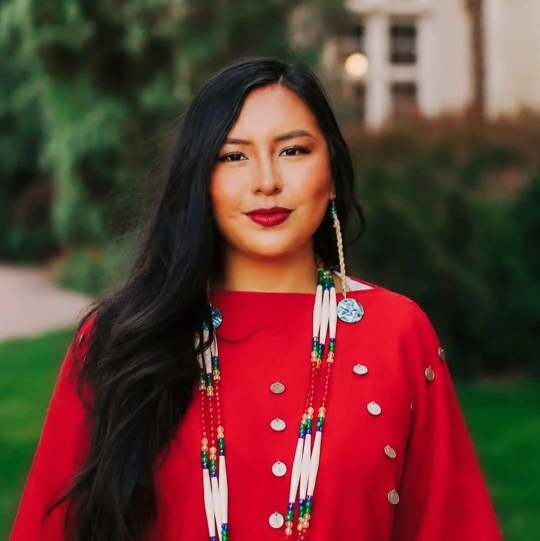 A woman with long black hair wearing a red shirt adorned with coin-like decorations and colorful beaded necklaces, standing outdoors with blurred greenery and house in the background.