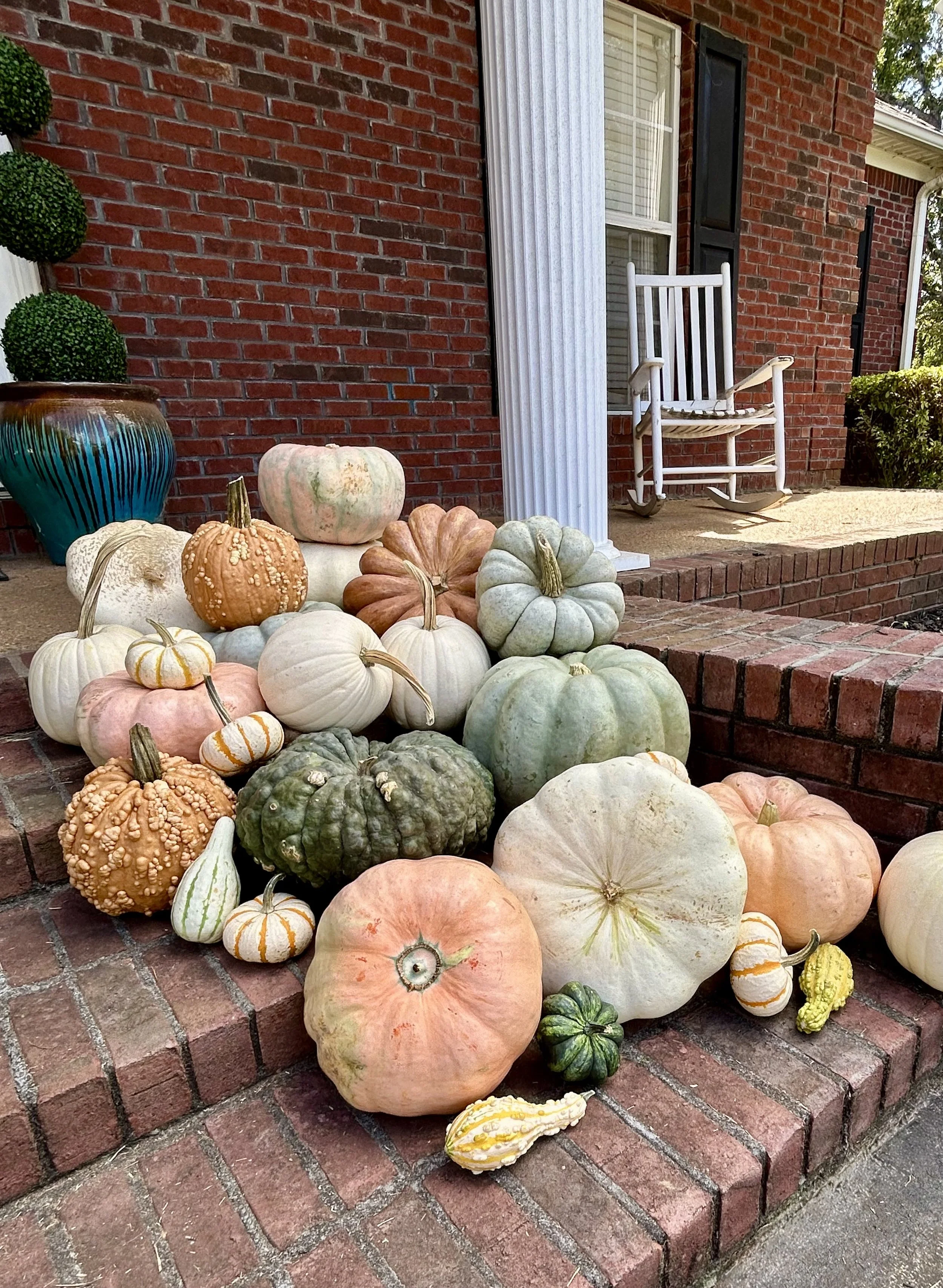Various pumpkins and gourds of different shapes, sizes, and colors are arranged on brick steps in front of a brick house with a window, white column, and white rocking chair.