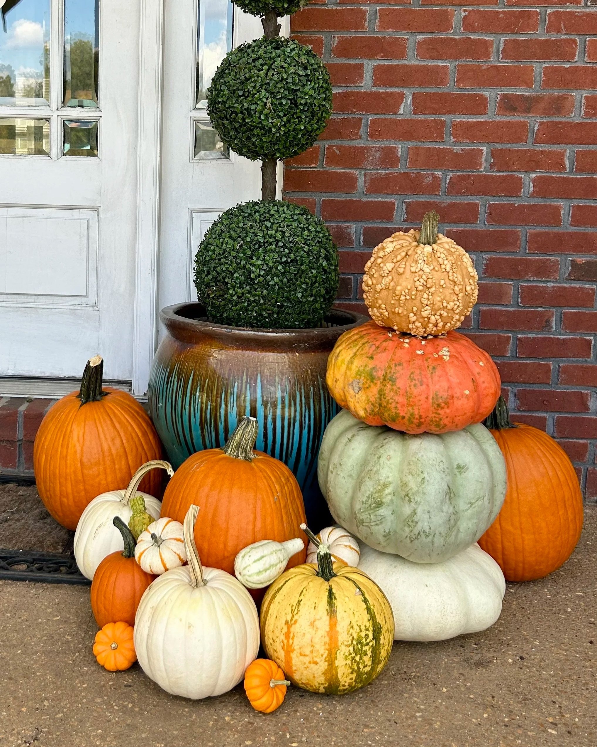 A stack of various pumpkins and gourds arranged outdoors against a brick wall and a white door. The pumpkins and gourds are in different sizes, shapes, and colors, including orange, white, green, and striped varieties. Two well-manicured topiary balls sit on a large decorative pot at the top of the stack.