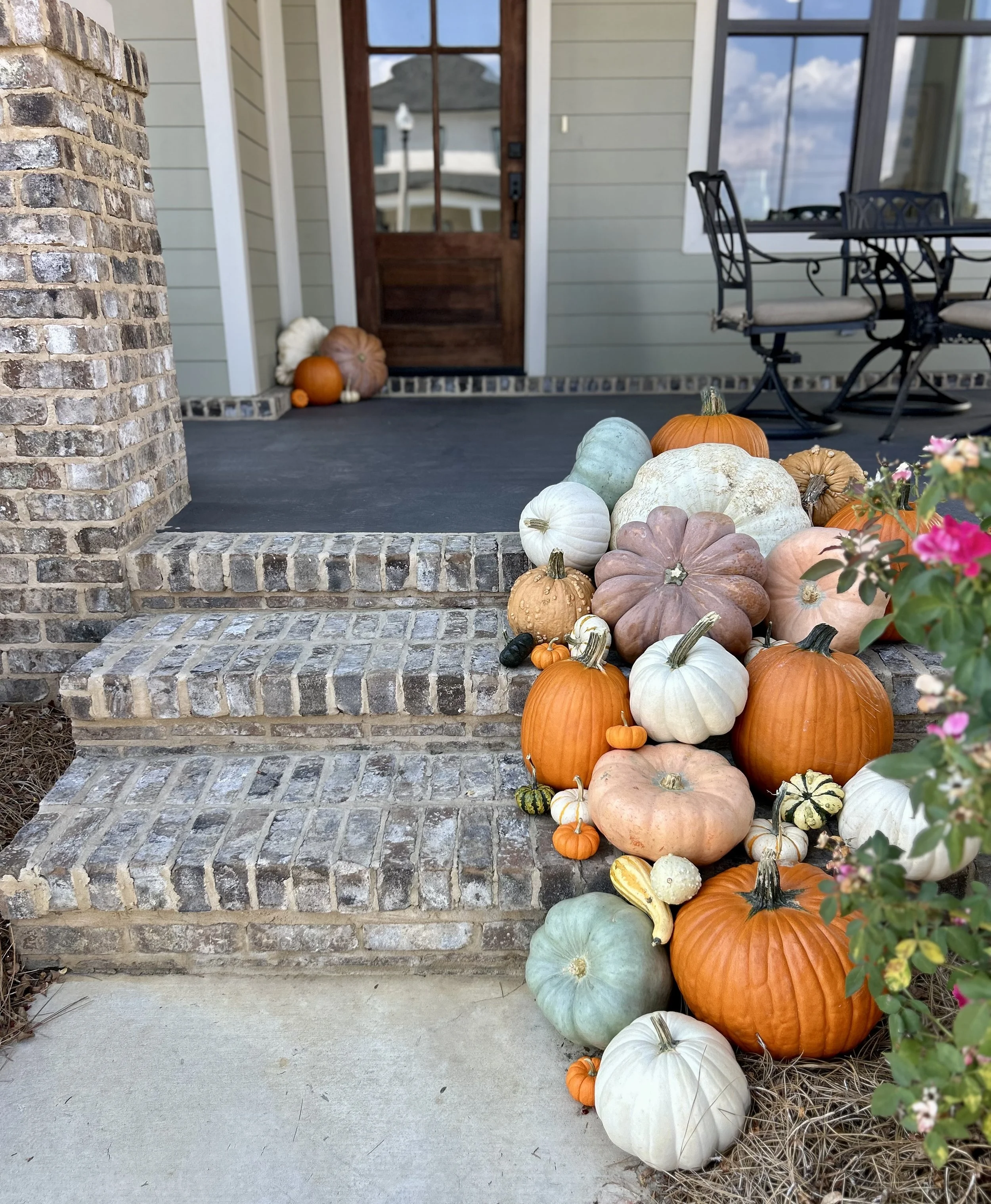 Fall pumpkins and gourds arranged on a step in front of a house porch with a brick column on the left, a wooden door in the center, and a window on the right.
