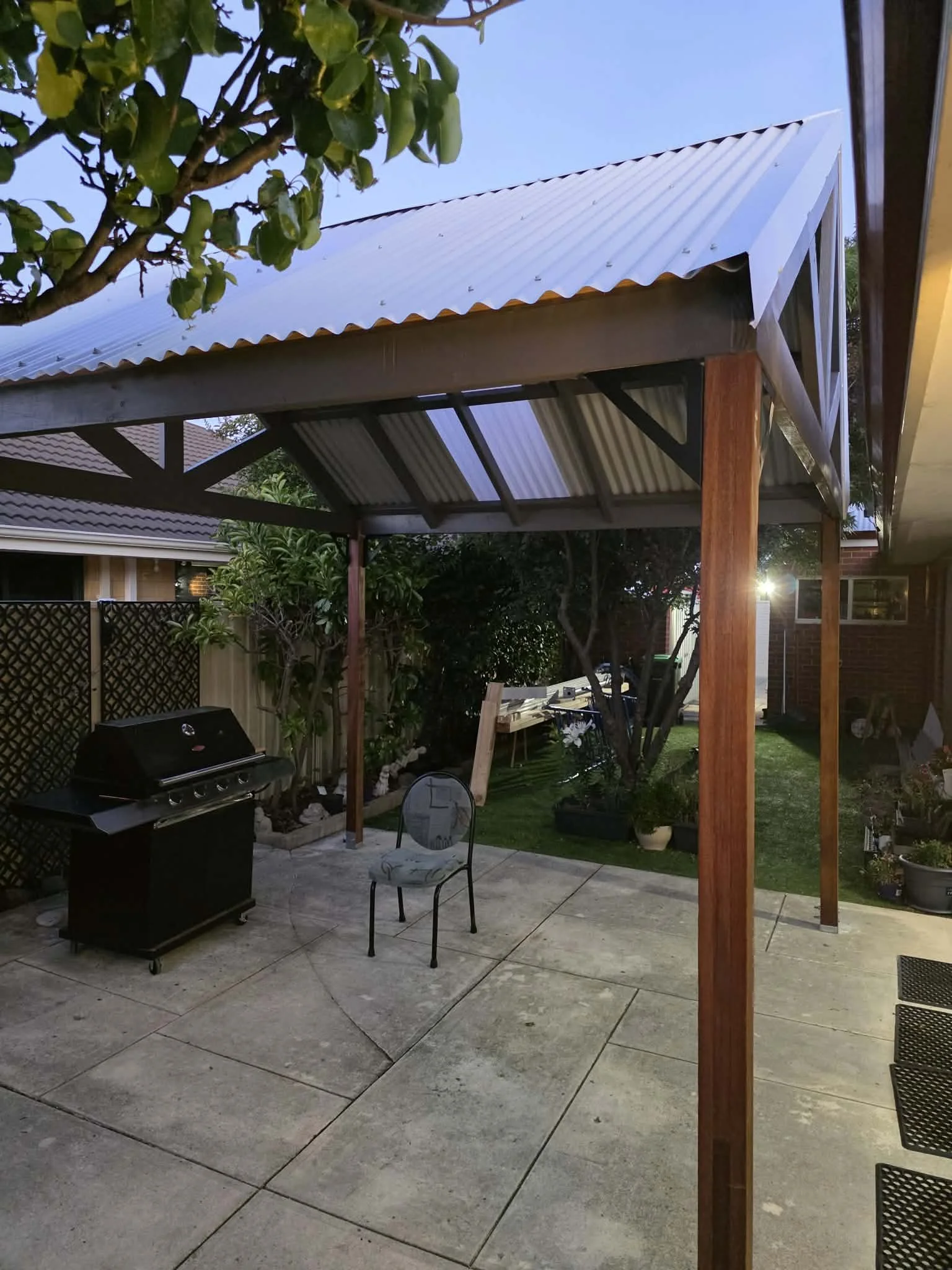 An outdoor patio area with a barbecue grill, a single chair with a decorative back, a tree, potted plants, and a covered structure with wooden and metal elements. The sky is clear and it appears to be evening.