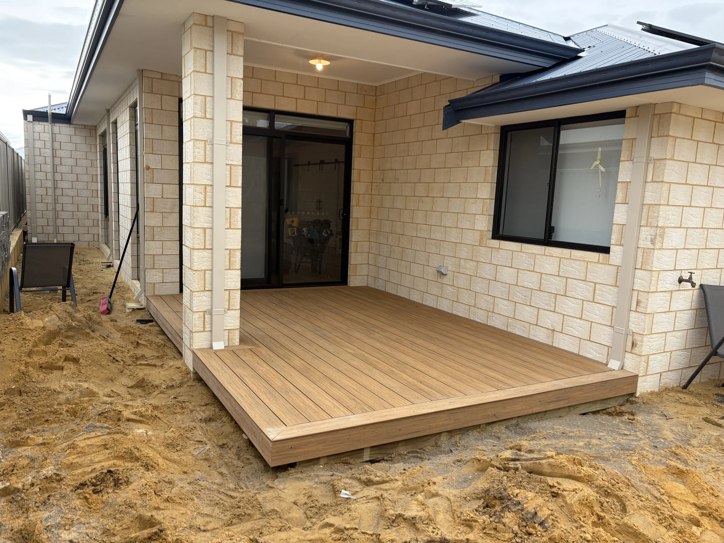 Back patio area with a wooden deck next to a house made of beige bricks, with black framed windows and sliding glass door, under a metal roof.