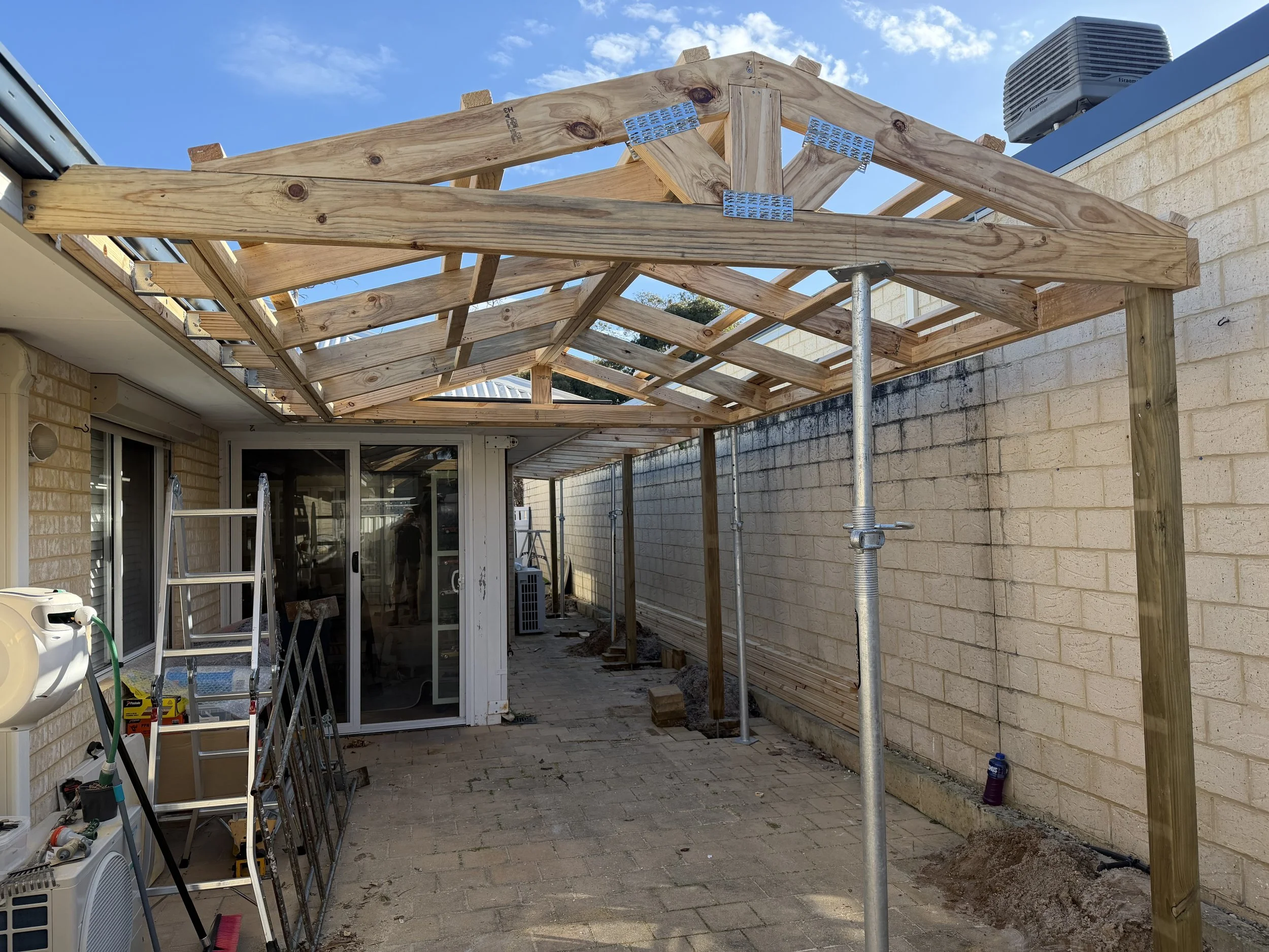 Construction site with a new wooden roof structure being built outside a house, scaffolding and tools visible on the ground.