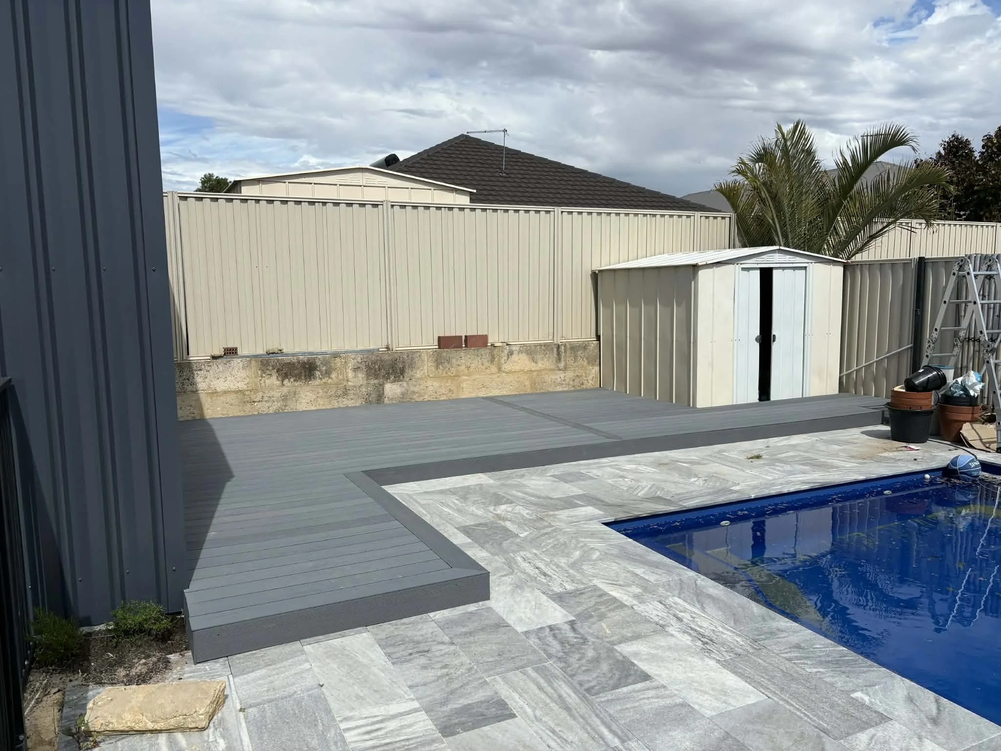 A backyard with a swimming pool, grey tiled deck, a beige fence, a small white shed, a ladder, and a large palm tree in the background on a partly cloudy day.