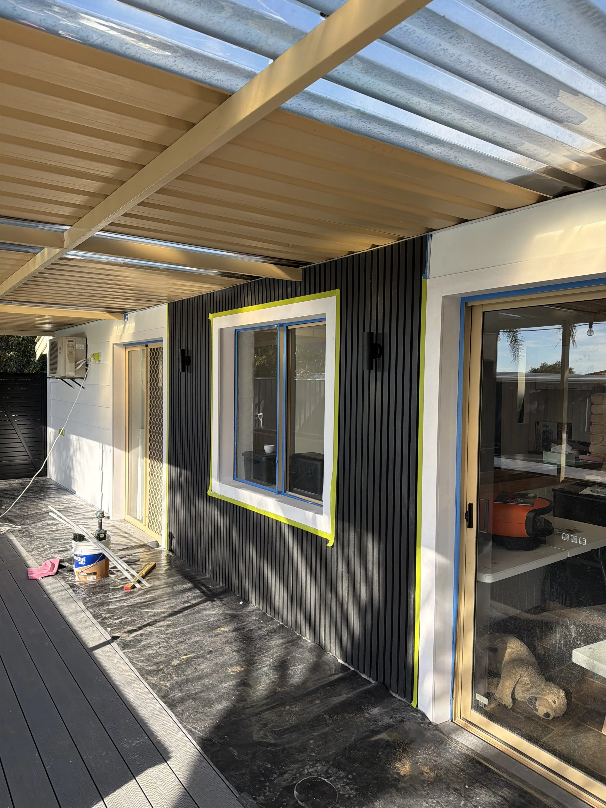 Exterior of a house under construction or renovation, with black siding, large windows, and a newly installed wooden ceiling; construction tools and supplies are on the ground.