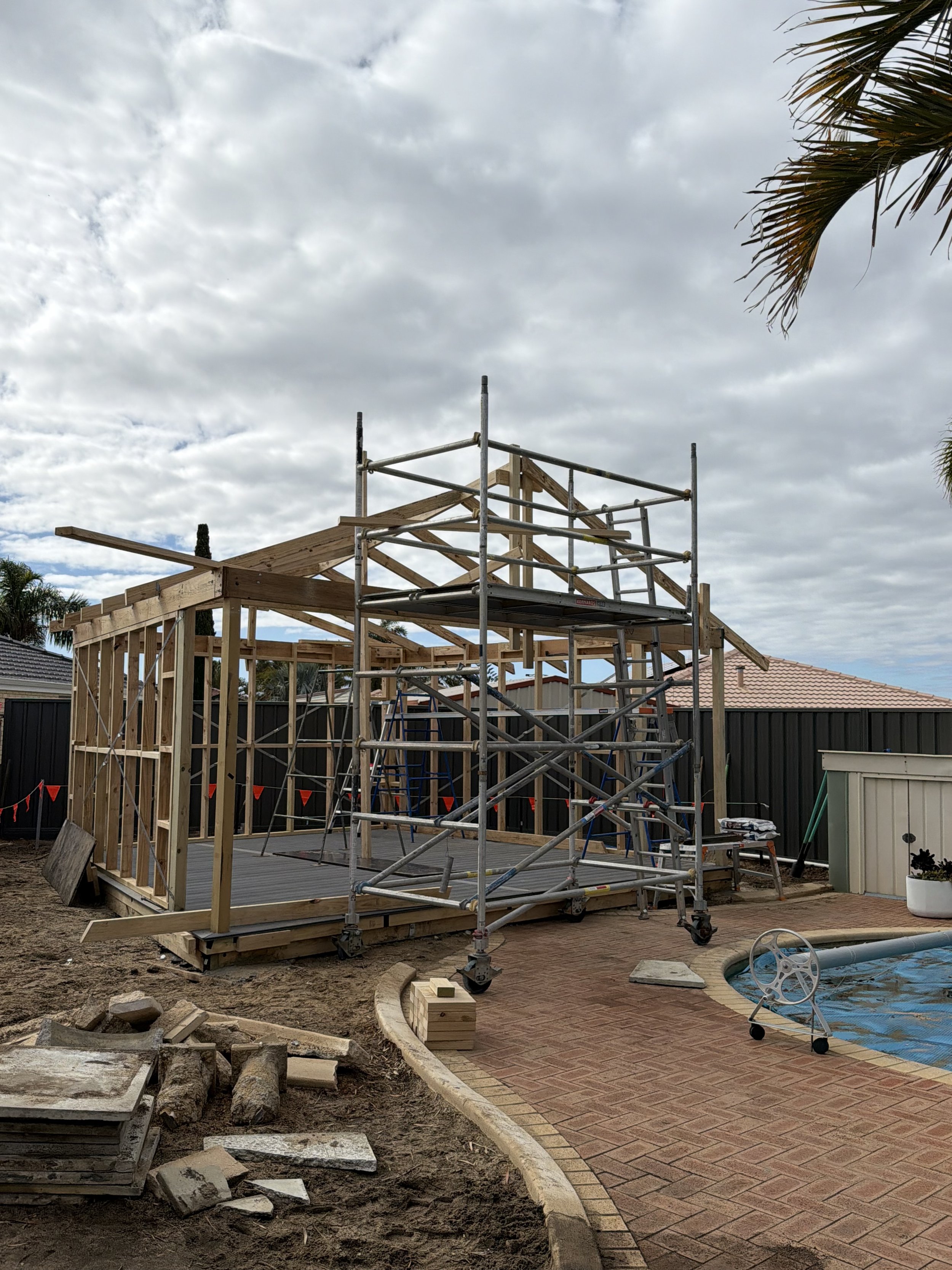 Construction site with a wooden framework, scaffolding, and a partially completed structure, alongside a paved pathway and construction materials on the ground under a cloudy sky.