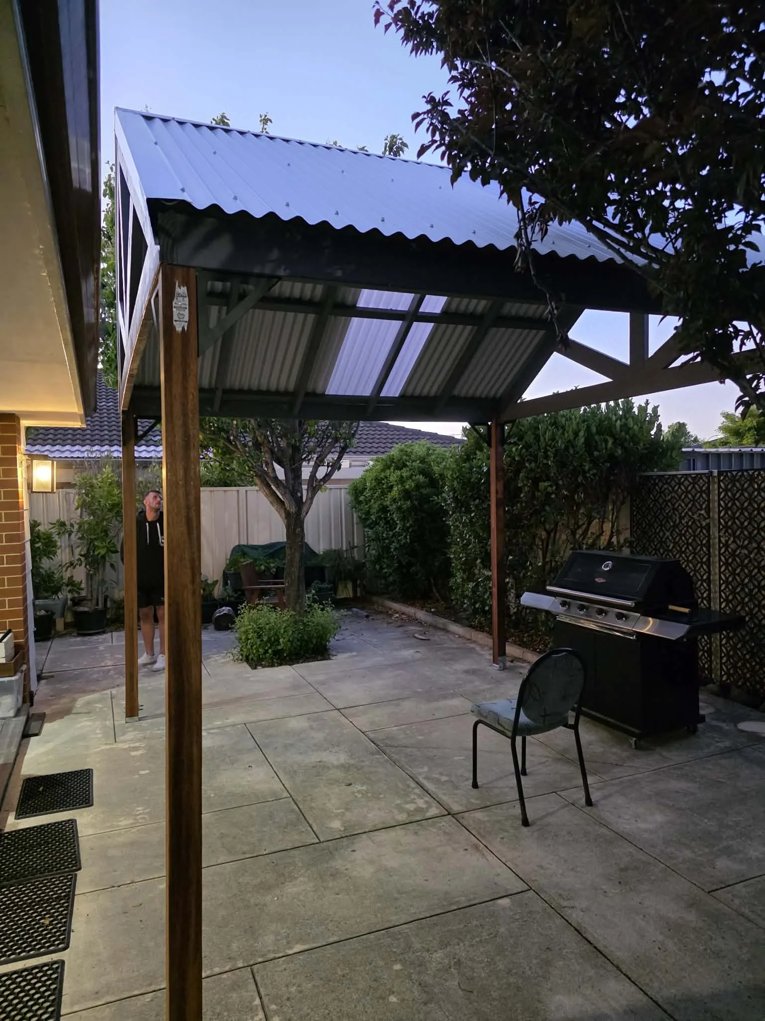 A backyard patio area with a covered pergola, trees, bushes, plants, a person standing near the house, and a grill.