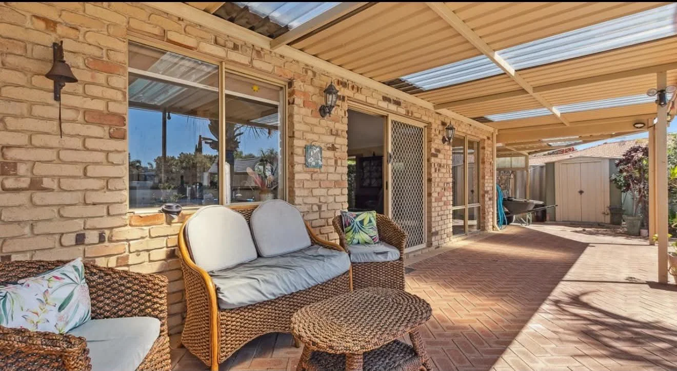 Outdoor patio with brick wall, wicker furniture with cushions, a small wicker table, sliding glass doors with security screen, wall-mounted lanterns, and a covered roof with wooden and metal beams.