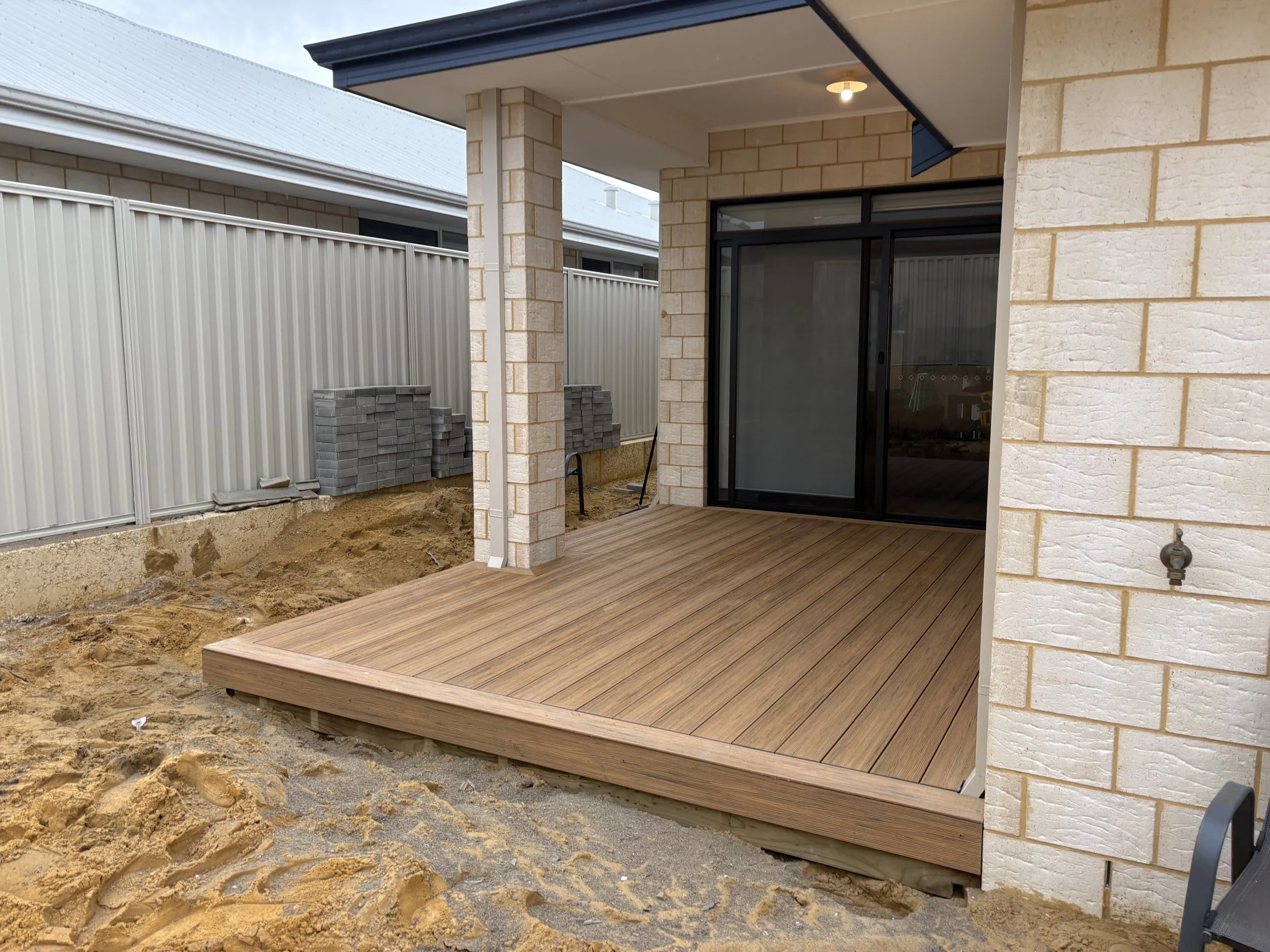 Newly constructed house with a wooden deck, brick walls, sliding glass door, and a construction site in the foreground with dirt and building materials.