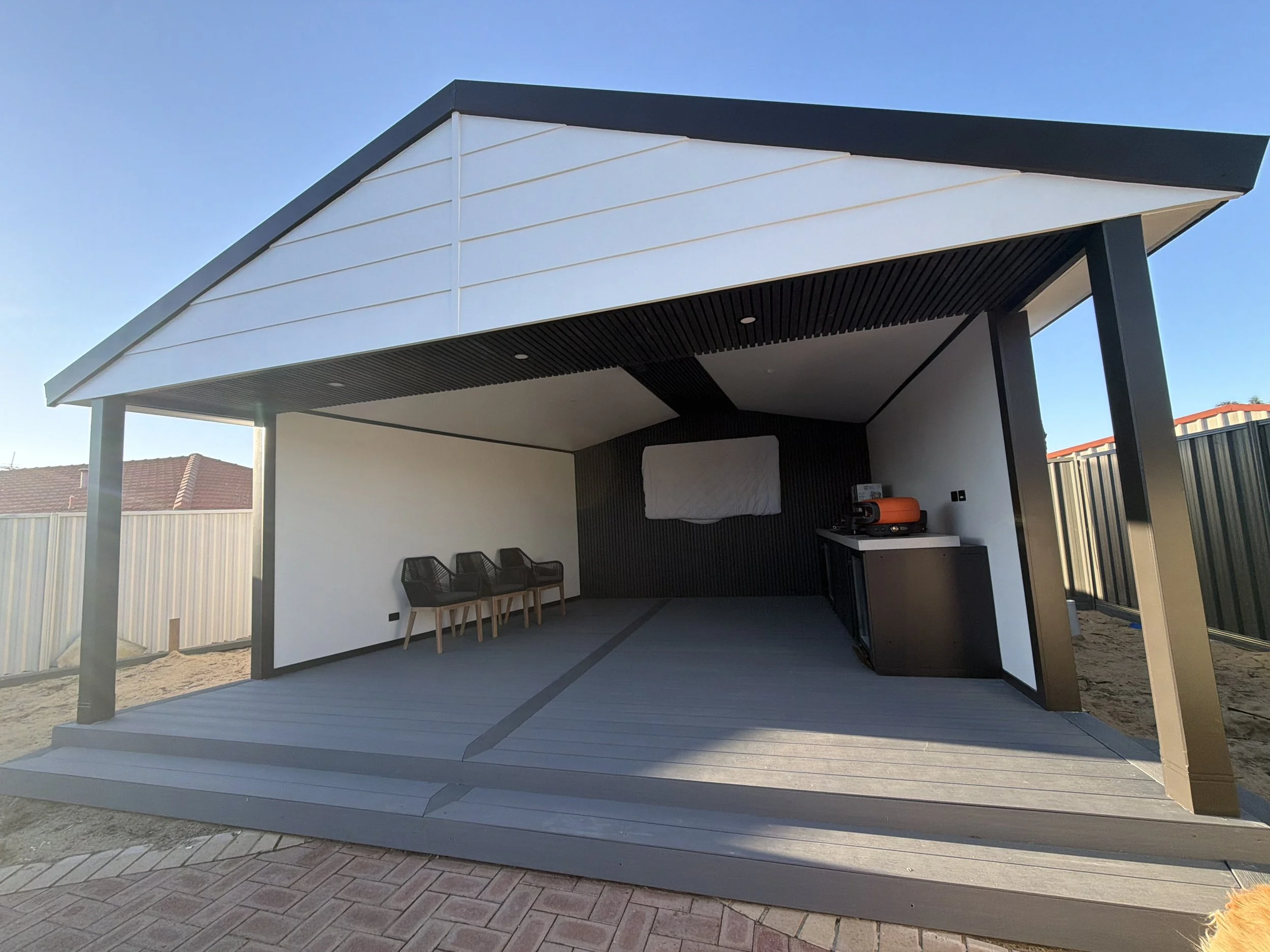 Modern outdoor covered patio with a grey deck, black support beams, and a white ceiling. There are three black chairs against the white wall and a small outdoor grill.
