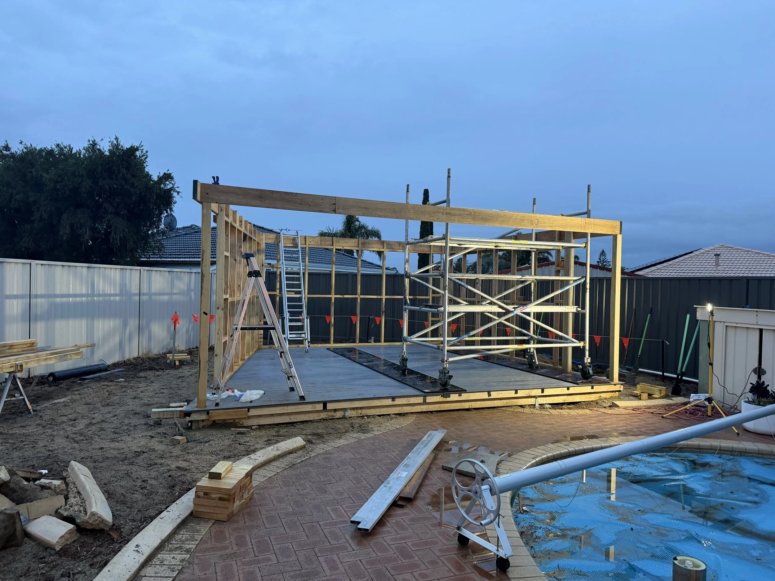 A backyard under construction with a wooden frame structure, scaffolding, a ladder, and building materials, with a house with a tiled roof and trees in the background under a partly cloudy sky.