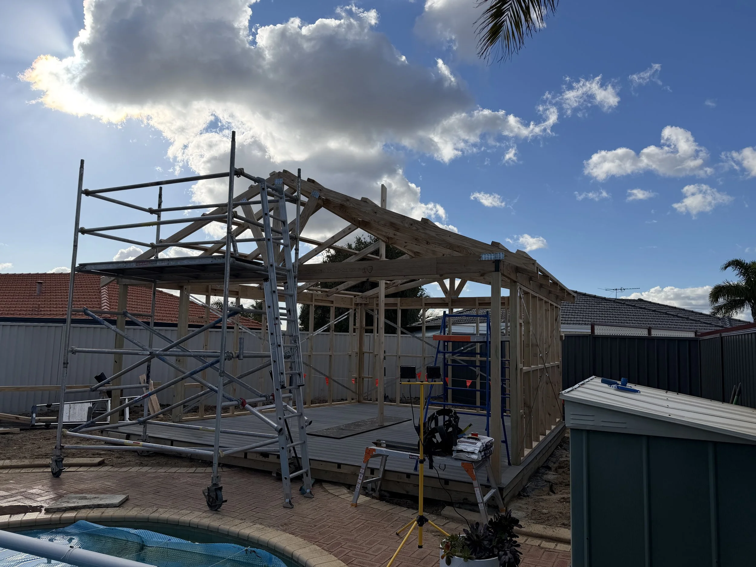 Construction site in backyard with wooden framework, scaffolding, construction tools, and equipment, under a blue sky with clouds.