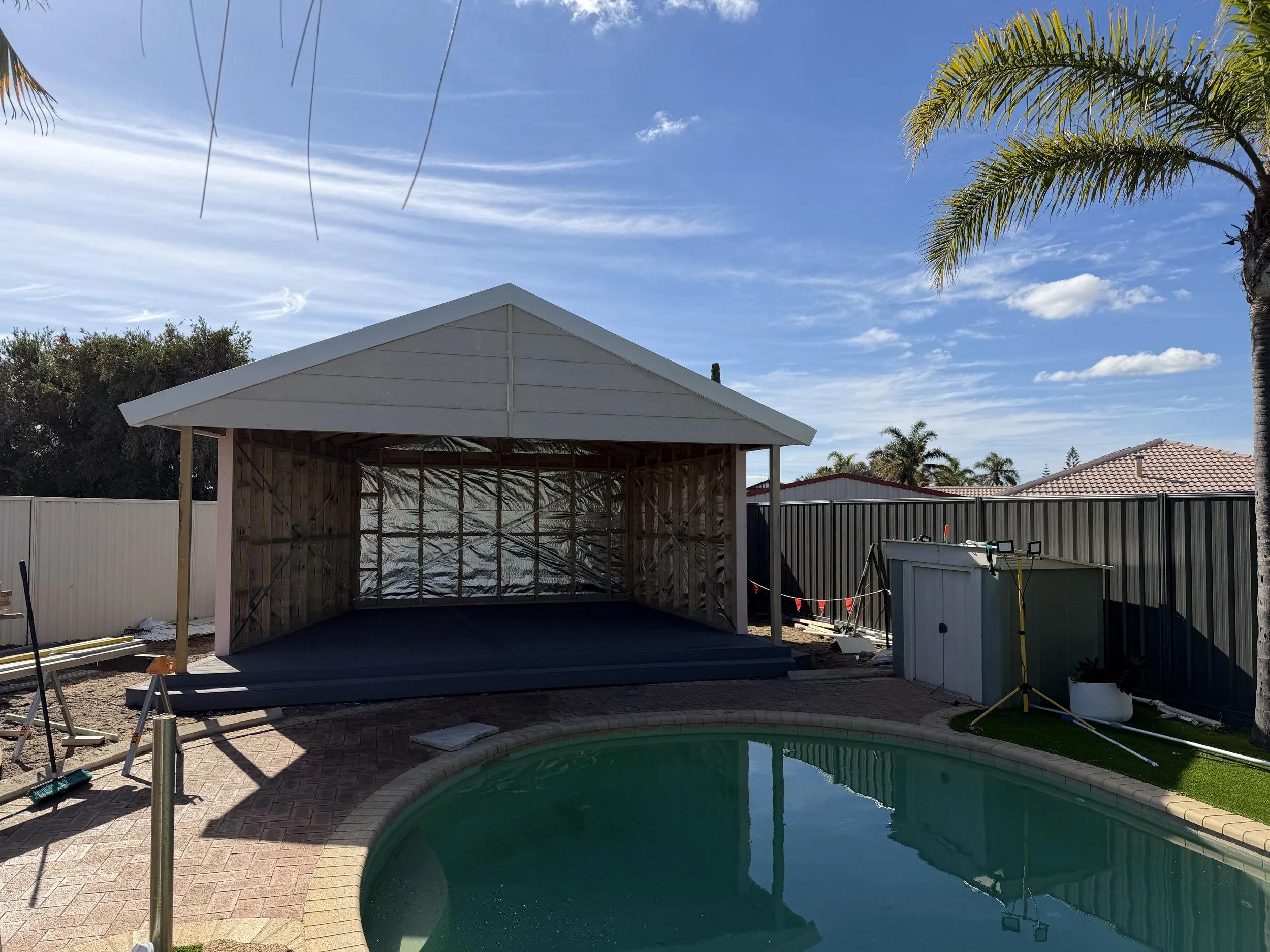 Backyard scene with a swimming pool in the foreground, an unfinished gazebo with a sloped roof frame in the middle, and a palm tree and neighboring rooftops under a blue sky with wispy clouds in the background.