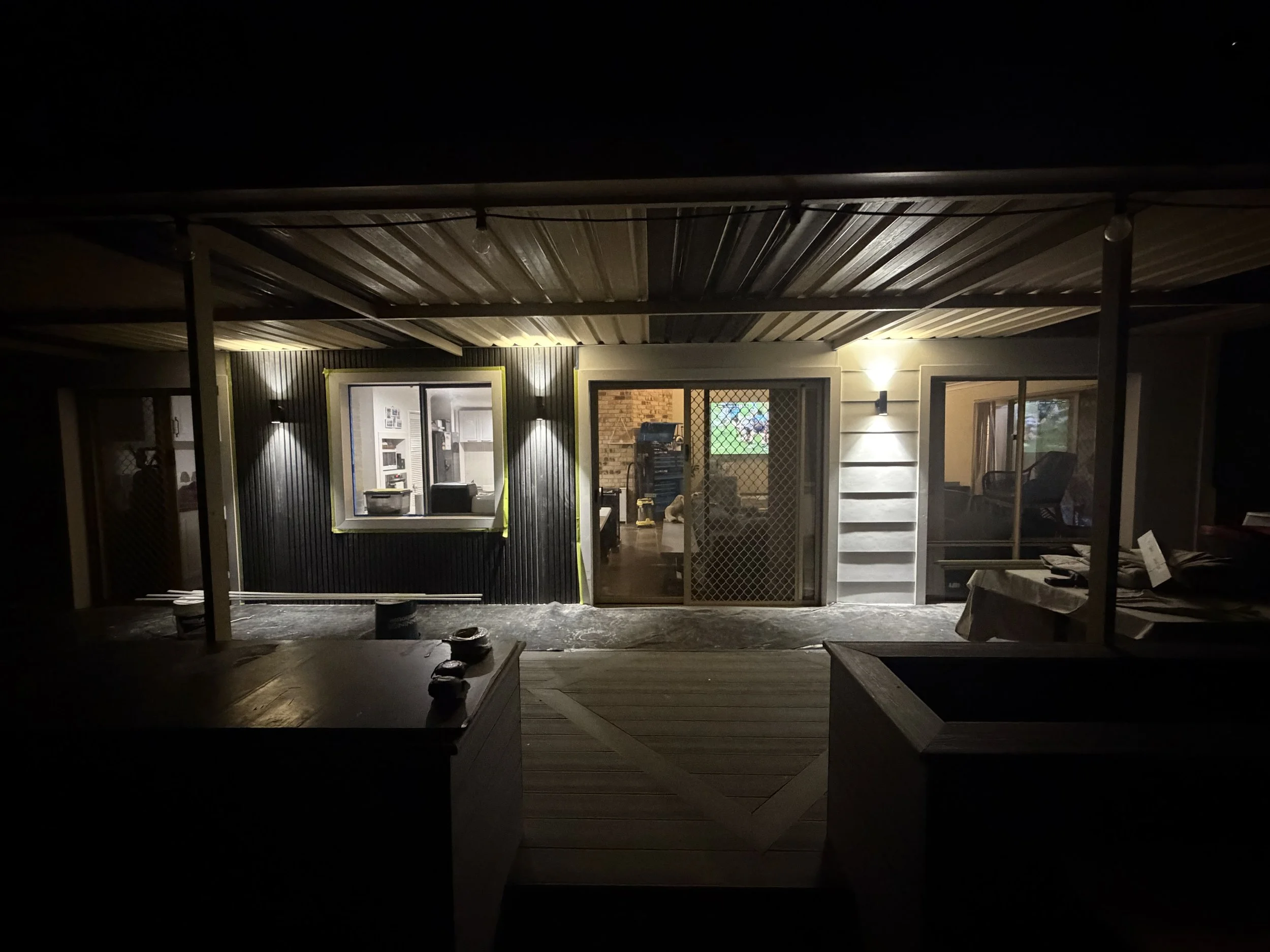 Nighttime view of a house under construction with a wooden deck, illuminated by interior and exterior lights, and unfinished roofing.