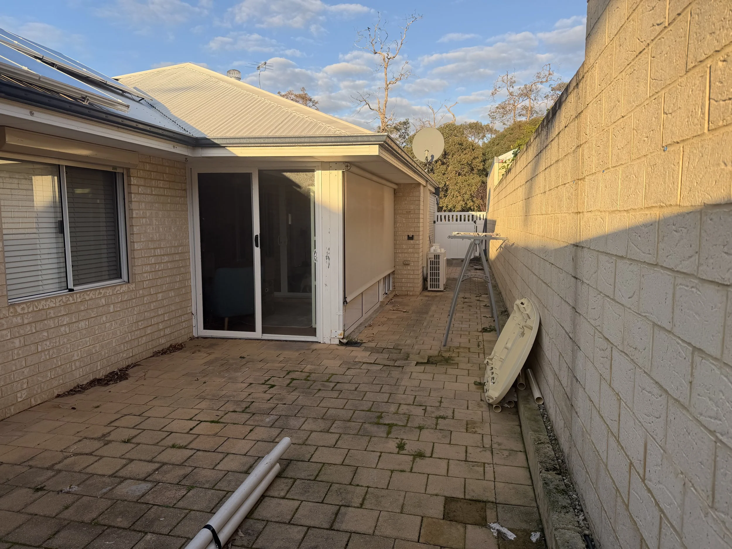 Backyard patio with brick flooring, beige brick house with white slide door and window, white fence, and stacked white pipes on the ground, with a partly cloudy blue sky and leafless trees in the background.
