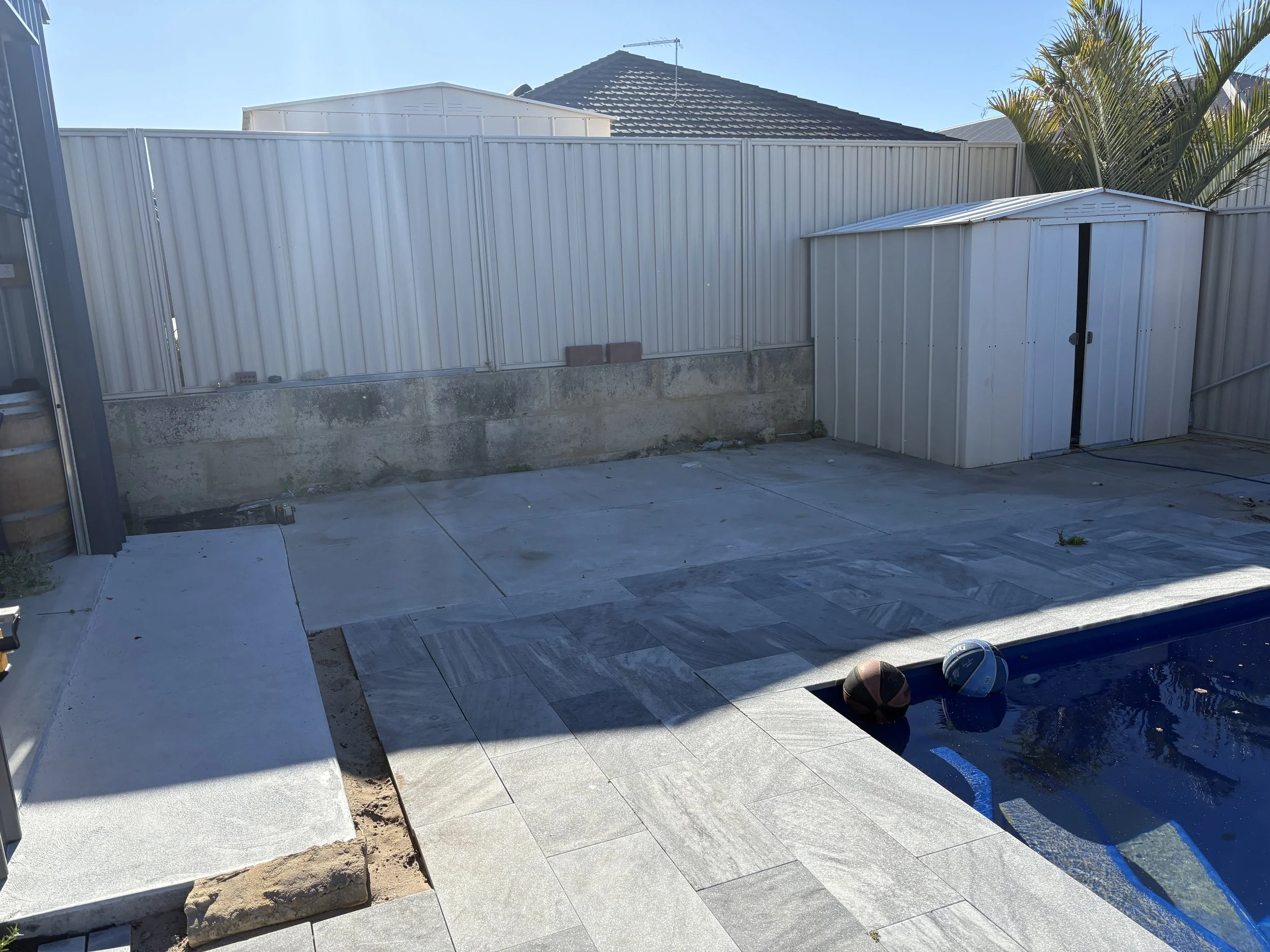 Backyard with a white metal fence, a small shed, and a swimming pool with floating toys; the patio is being tiled.