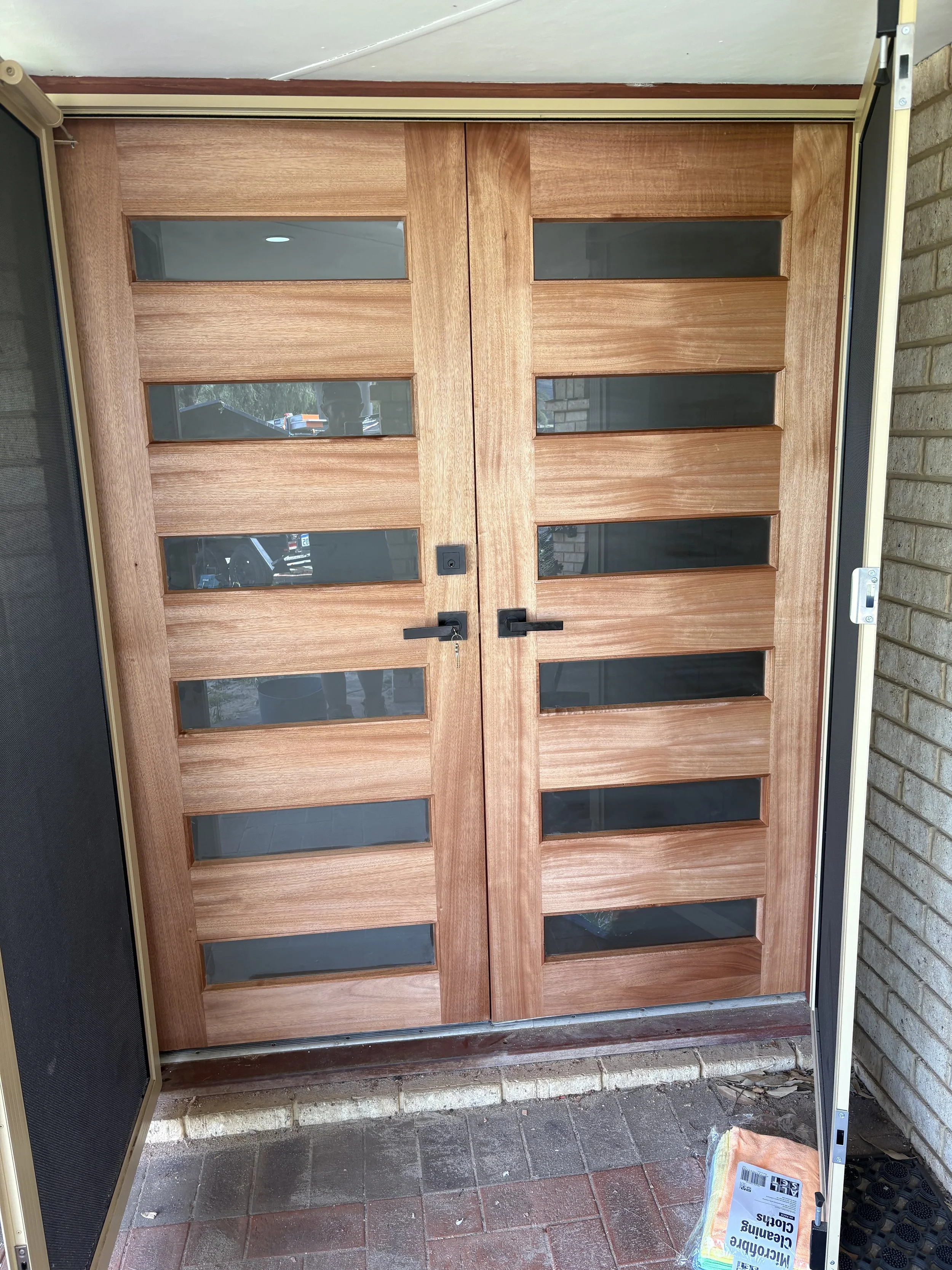 A pair of closed wooden doors with glass panels at the top, set in a brick wall. The doors have black handles and a lock; the left door has a small chain lock. There is a black-screened panel on the left side and a package on the ground near the bottom right.