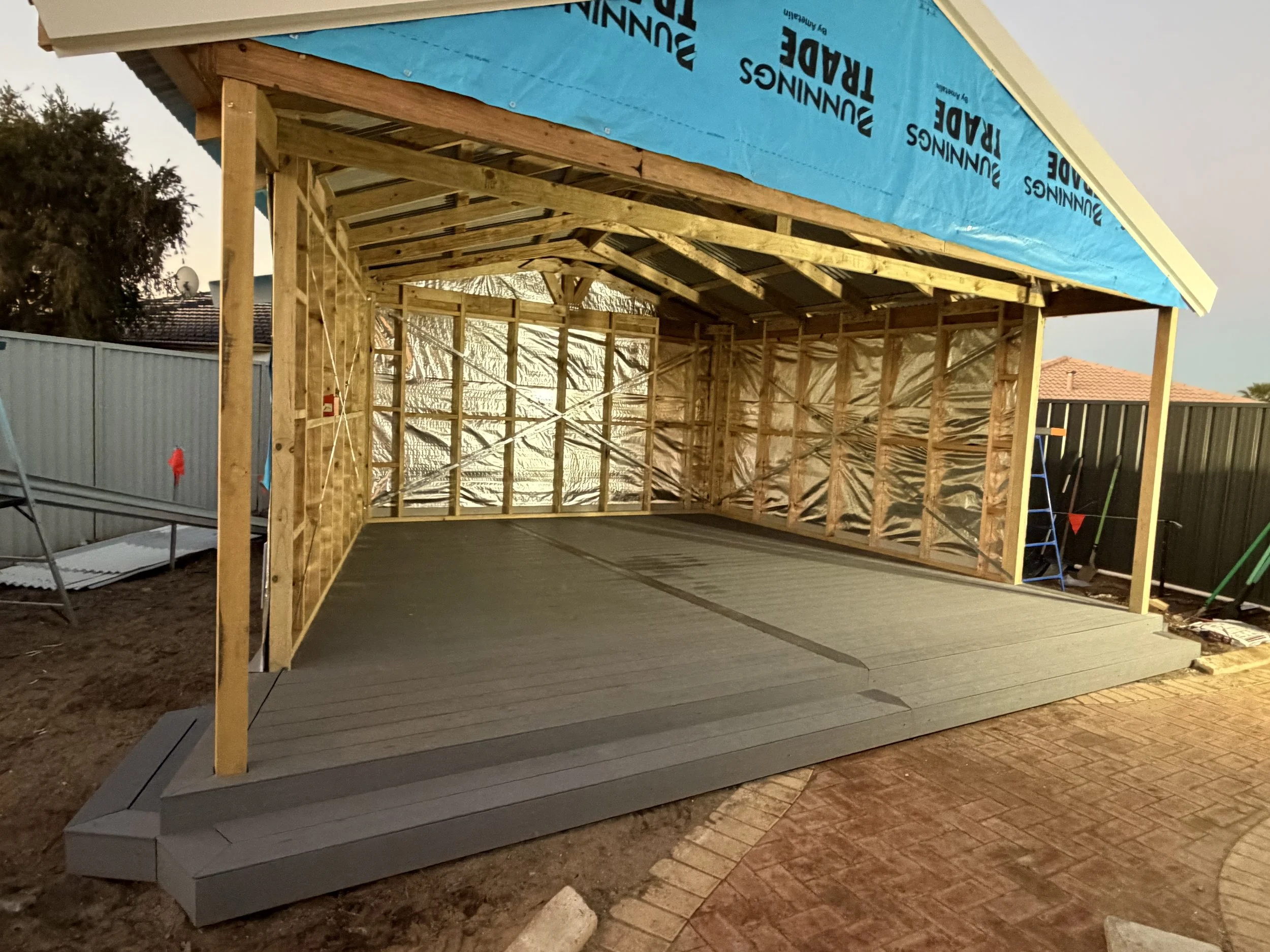 A partially constructed outdoor structure with a wooden frame, gray flooring, and a blue tarp covering the roof, situated in a backyard with fencing and a tile roof house in the background.
