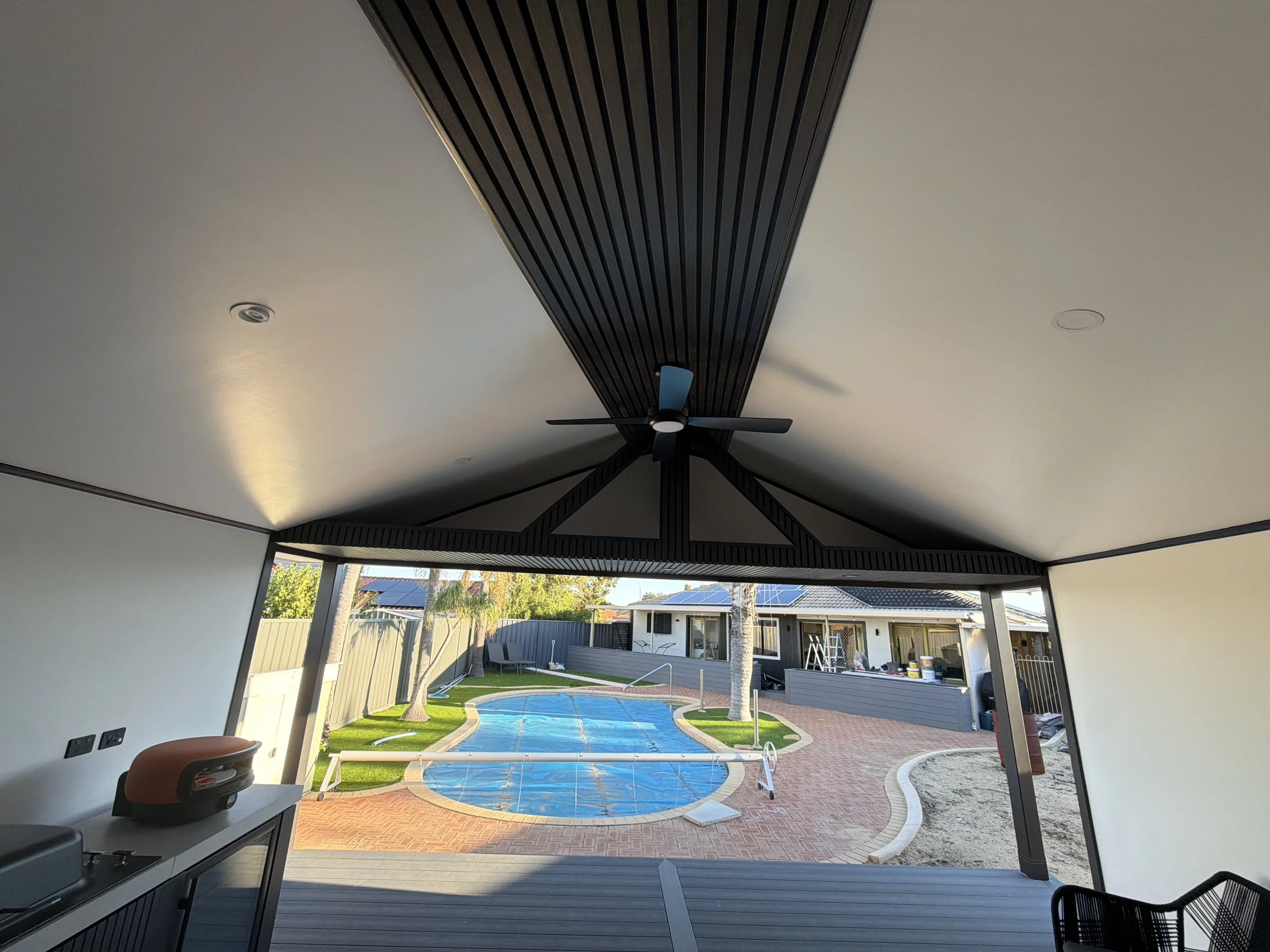 Looking through a covered outdoor patio to a backyard with a swimming pool, lawn, trees, and a house with solar panels.
