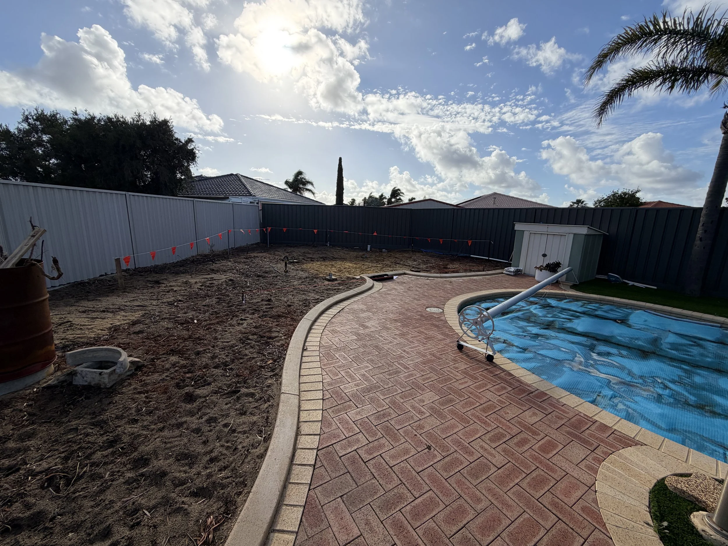 Backyard with a paved brick walkway, a covered pool, a tall dark fence, and a patch of dirt with construction materials, under a partly cloudy sky with sunlight.