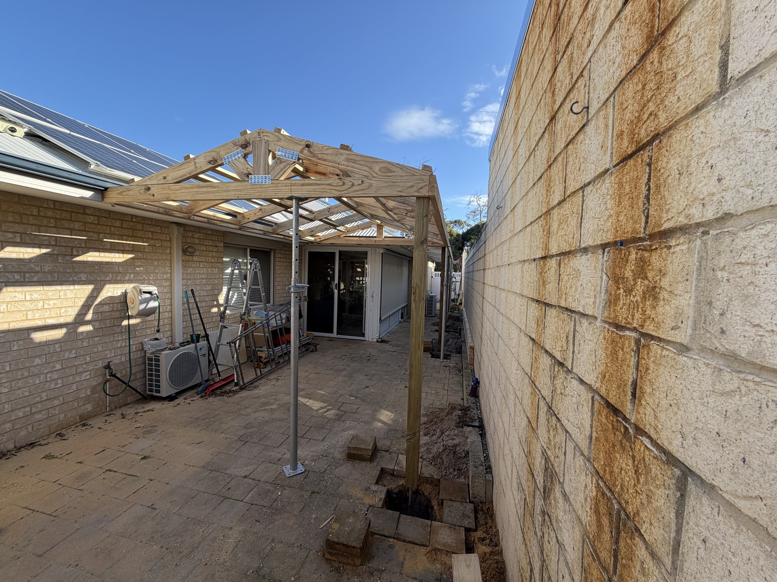 A backyard construction scene showing a wooden pergola frame being built, with scaffolding, ladders, and construction tools nearby. A brick house wall with solar panels and a stone wall are visible.