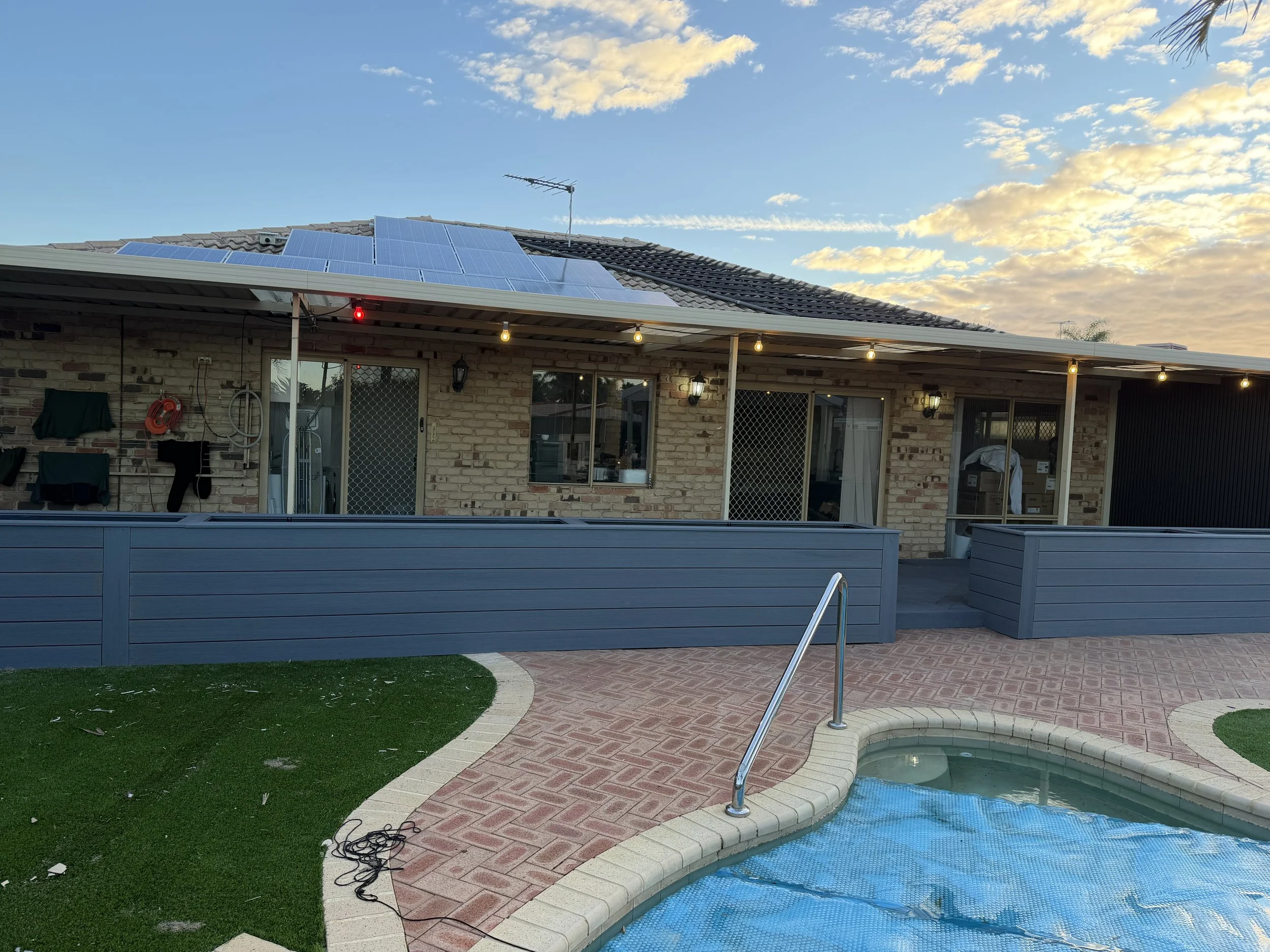 Backyard area with a swimming pool cover, brick pathway, artificial grass, and a house with solar panels on the roof under a partly cloudy sky at sunset.