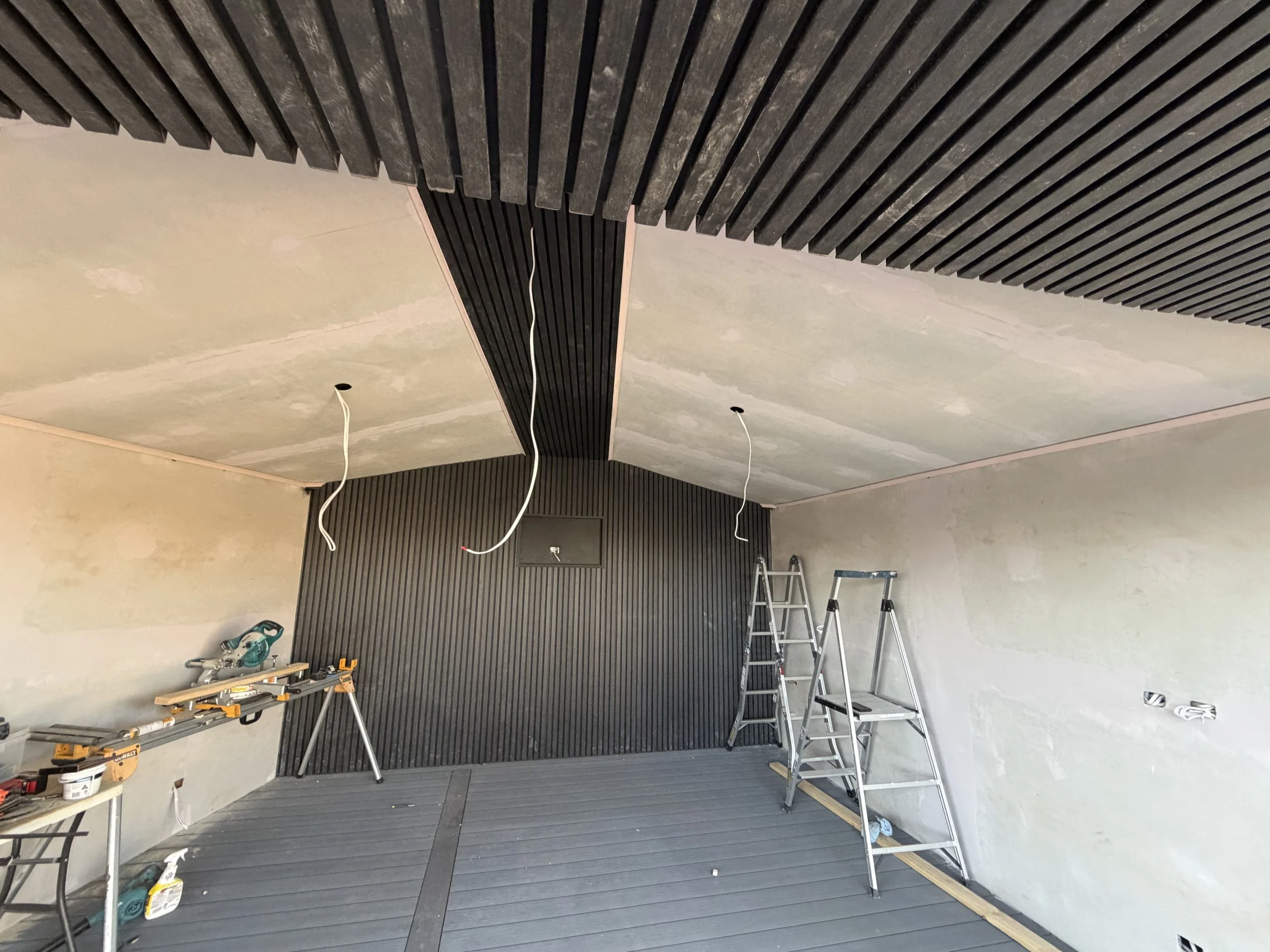 Room under renovation with ladders, tools, and black wall paneling being installed on the ceiling and wall.