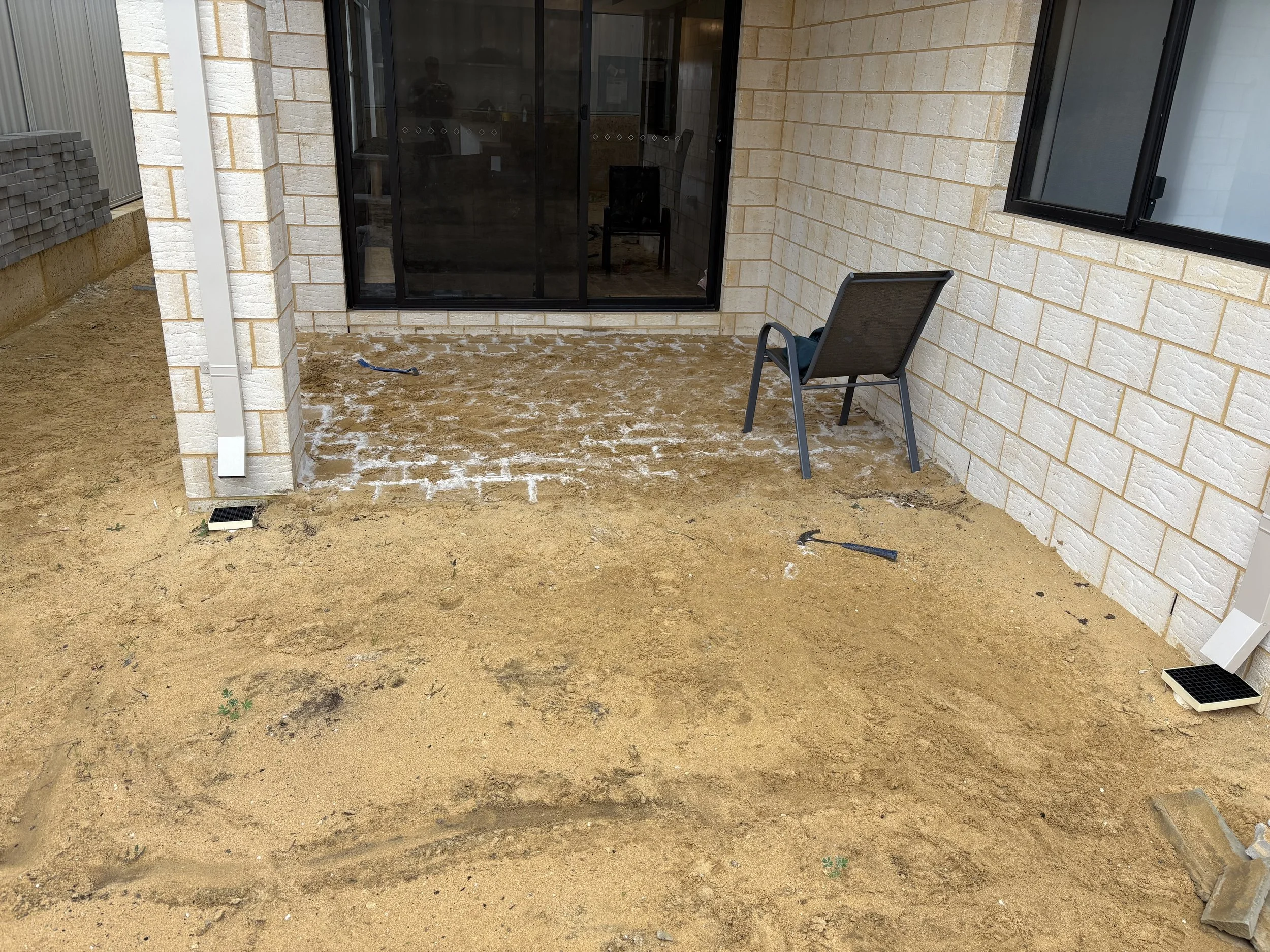 A backyard patio under construction with a dirt ground and sand scattered, enclosed by brick and siding walls, a black sliding glass door, a single chair, and a window.