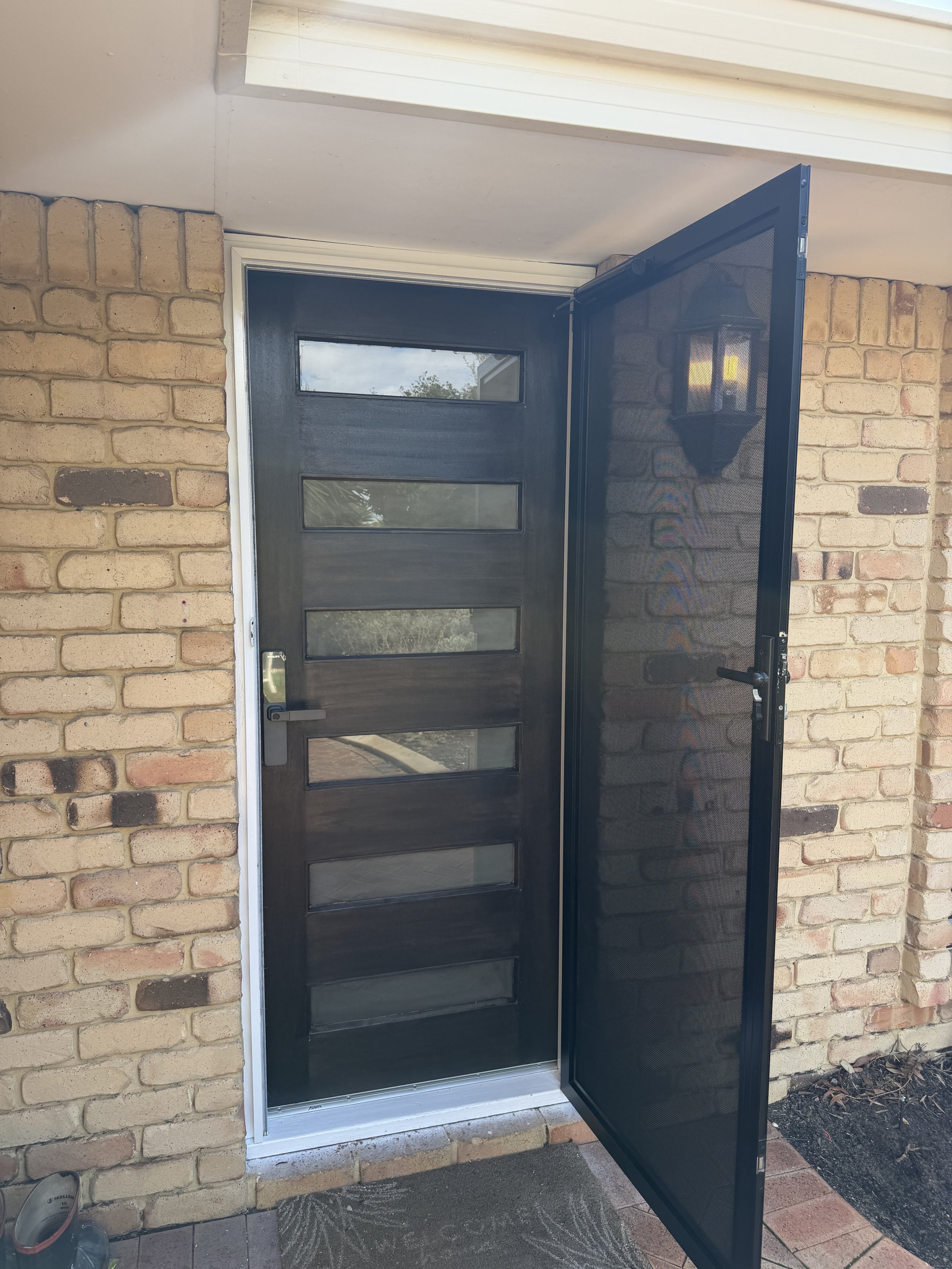 Front door of a house with a black security screen door partially open, brick wall on either side, and a small potted plant on the ground in front.
