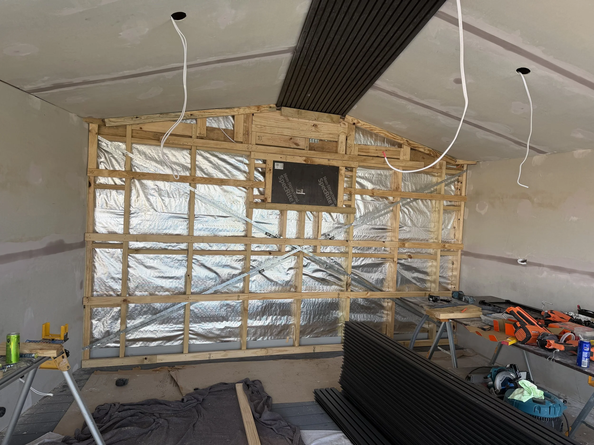 Construction room with wooden framing and soundproofing panels installed on the wall, tools and materials scattered around, including a drill, black panels, and wiring, in the process of being built or renovated.