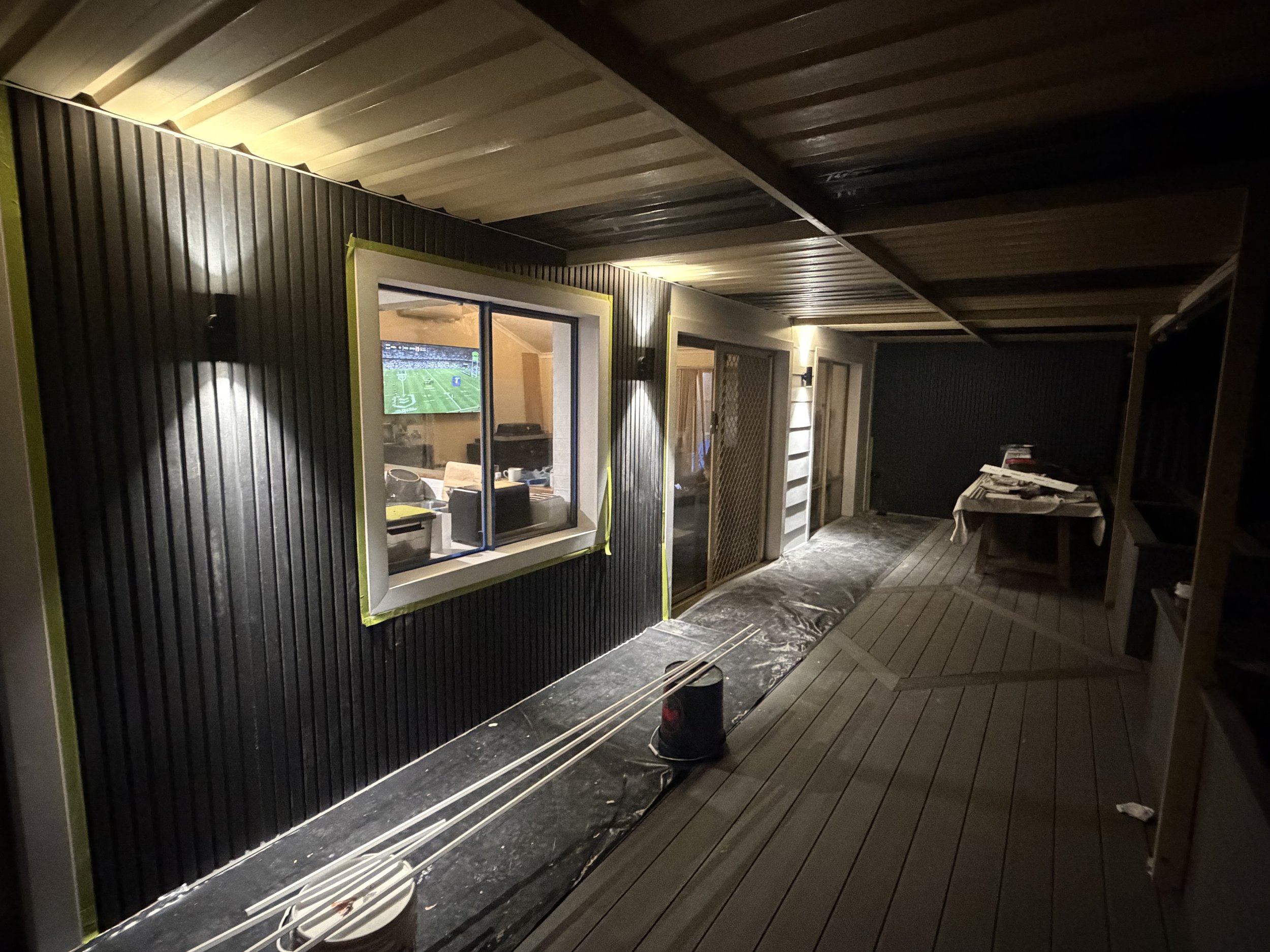 A covered porch with black and wood-paneled walls, a window showing a television with a football game, and various household items. There is a table with a tablecloth and some objects on it, a speaker, and a bucket on the floor.