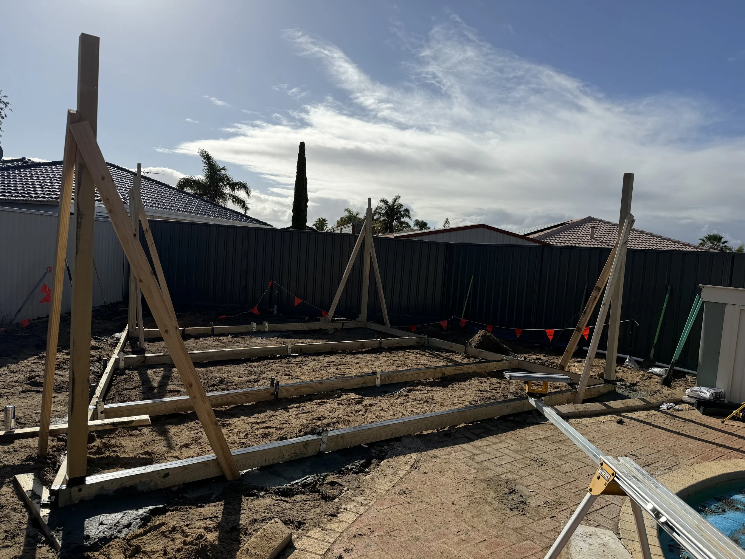 Construction site with wooden framing for a new building, fenced backyard, and cloudy sky.