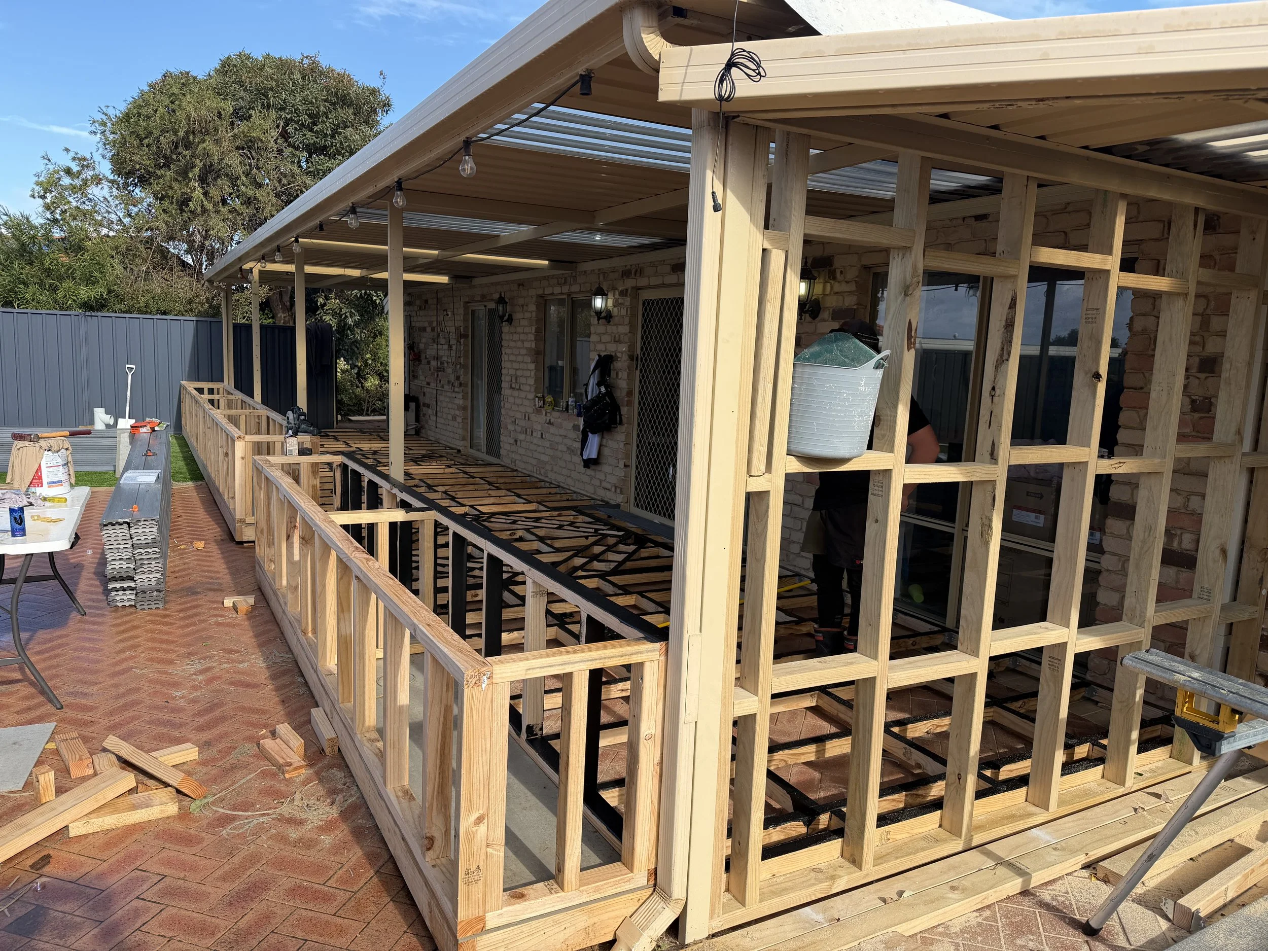 Construction of a wooden deck with framing, partially built outside a brick house with a sliding glass door and a screen door, under a roof supported by wooden posts, with construction tools and materials on the ground.