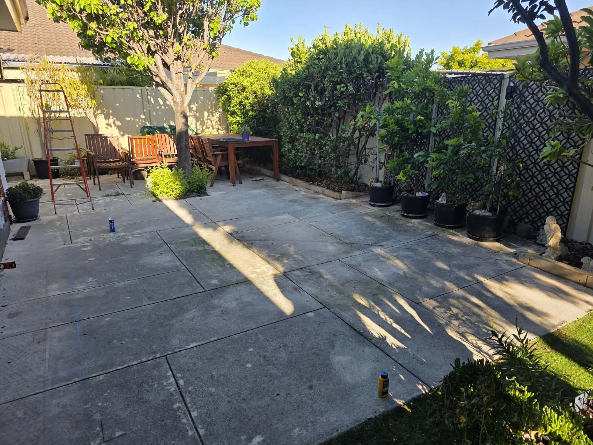 A backyard patio with outdoor furniture, potted plants, and garden tools, surrounded by trees, shrubs, and a private fence.