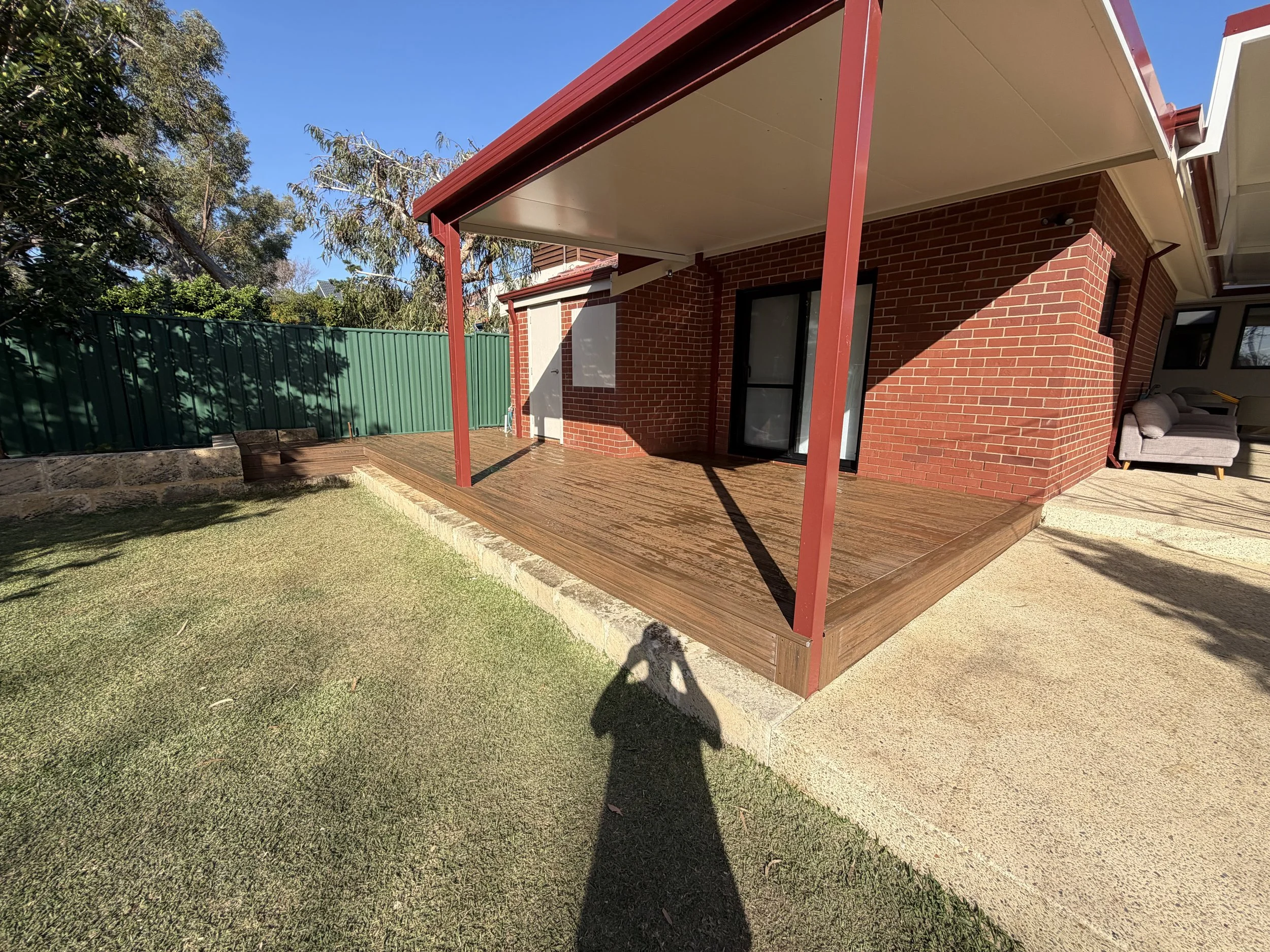 Backyard patio with wooden deck, brick house wall with sliding glass door, outdoor sofa, green fence, trees, and clear blue sky.