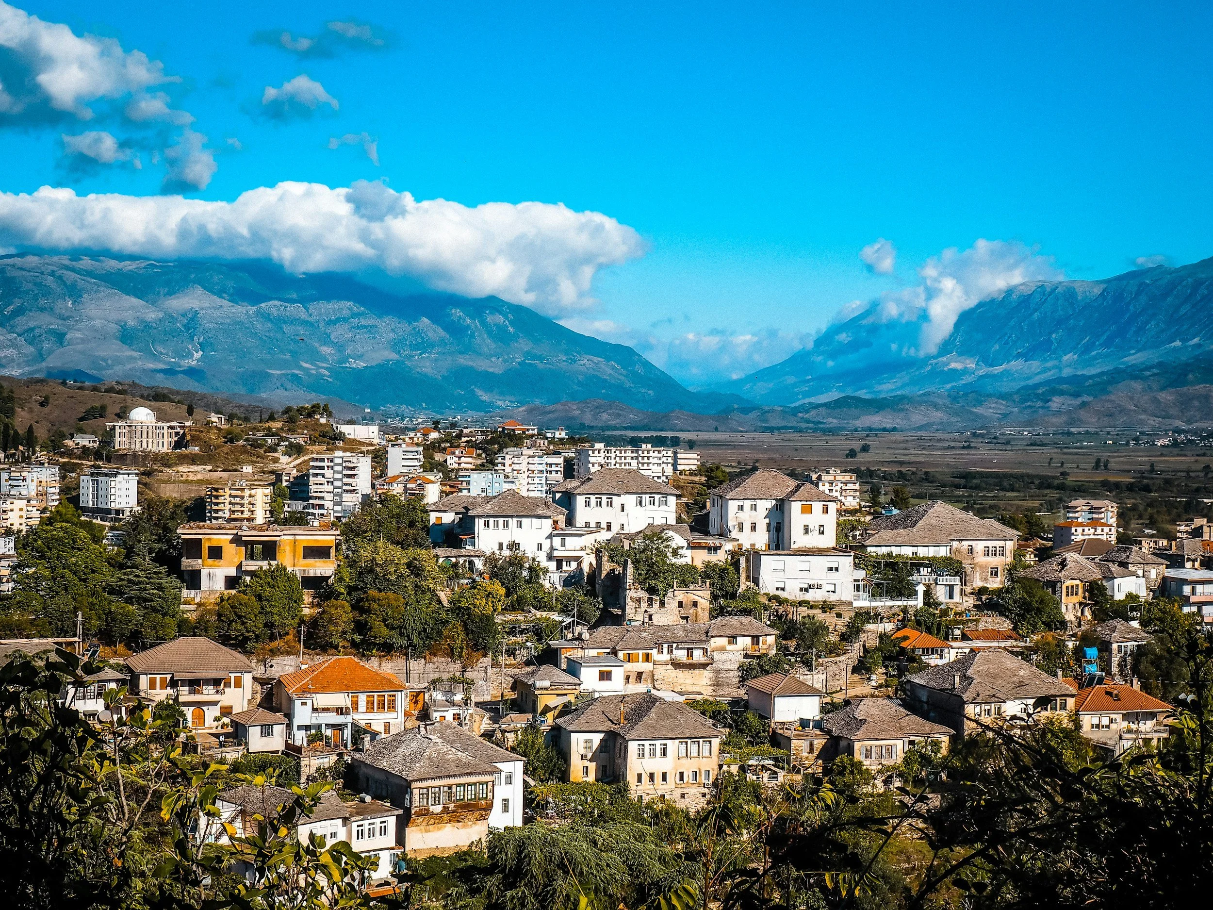 View of a mountain range in the background with a town in the foreground, featuring white buildings with orange and gray roofs, under a partly cloudy sky.