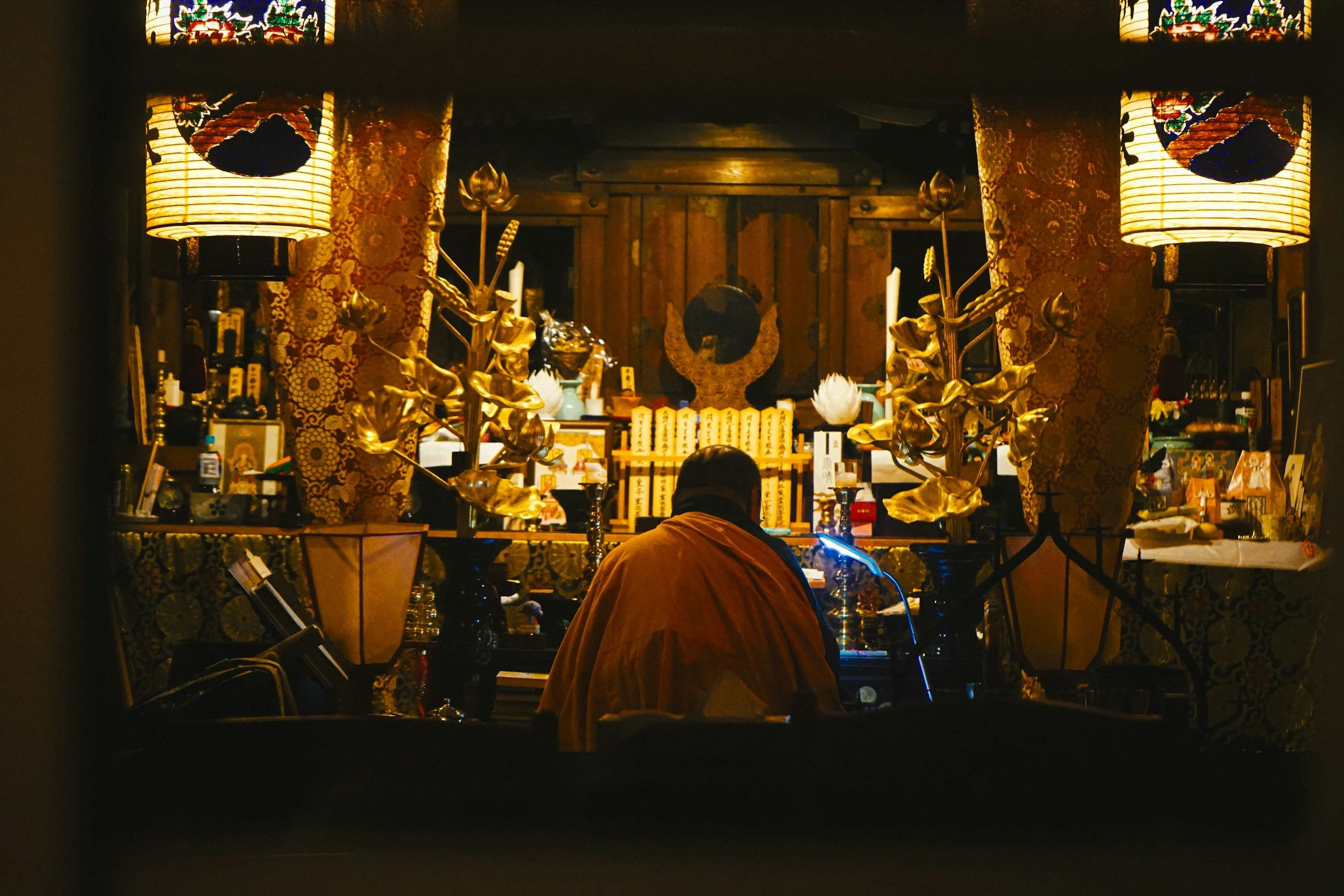 View through a small window of a traditional Japanese altar room decorated with gold statues, ornate lanterns, and intricate embroidered fabric. The person in the foreground is wearing an orange garment and seated facing the altar.